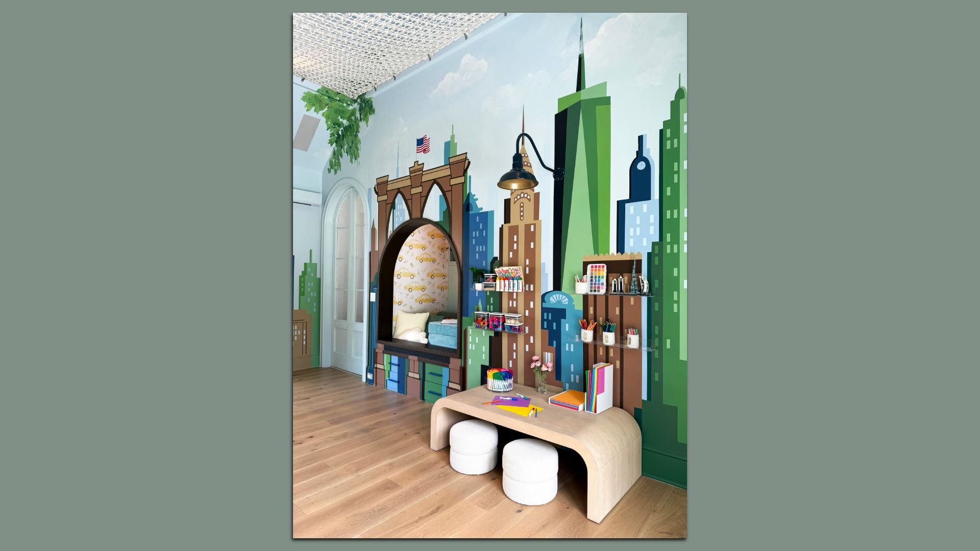 Colorful interior wall mural of a city skyline with tall buildings, arches and a hanging lamp. There's also a reading nook with taxicab wallpaper, shelves of art supplies, a curved wooden bench, round white stools and a table with colored papers.
