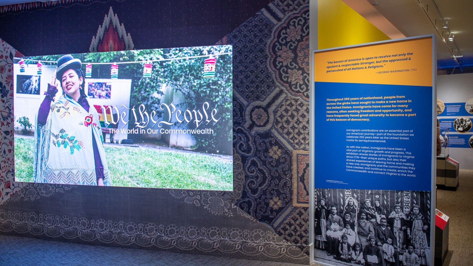 Exhibit with a large screen showing a smiling woman in a hat and poncho; the title "We the People" is visible, with a blue/yellow info panel and a vintage group photo beneath.