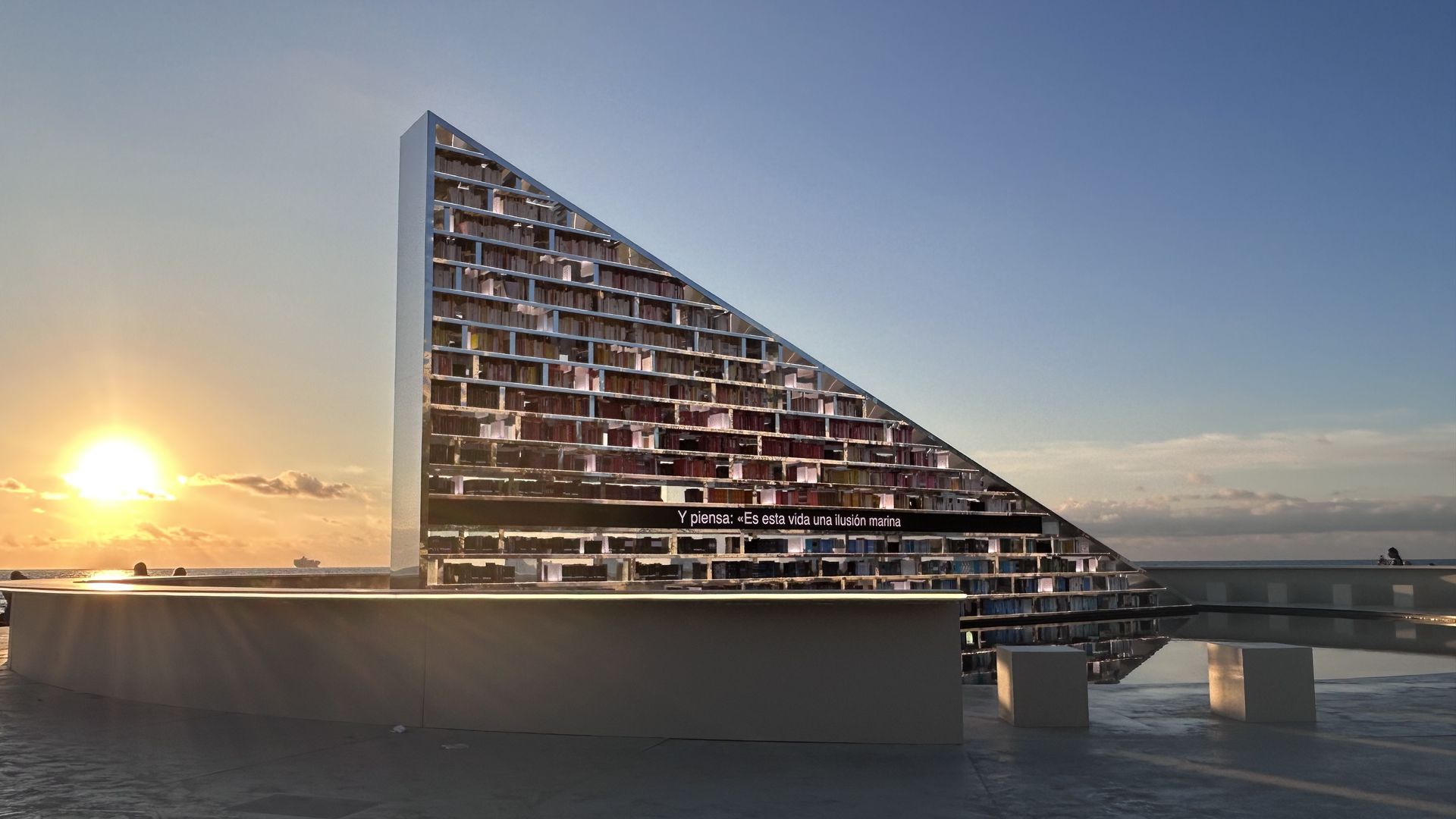 Sunset by a triangular mirrored monument with Spanish text reflecting the golden sun and blue sky over a calm sea.
