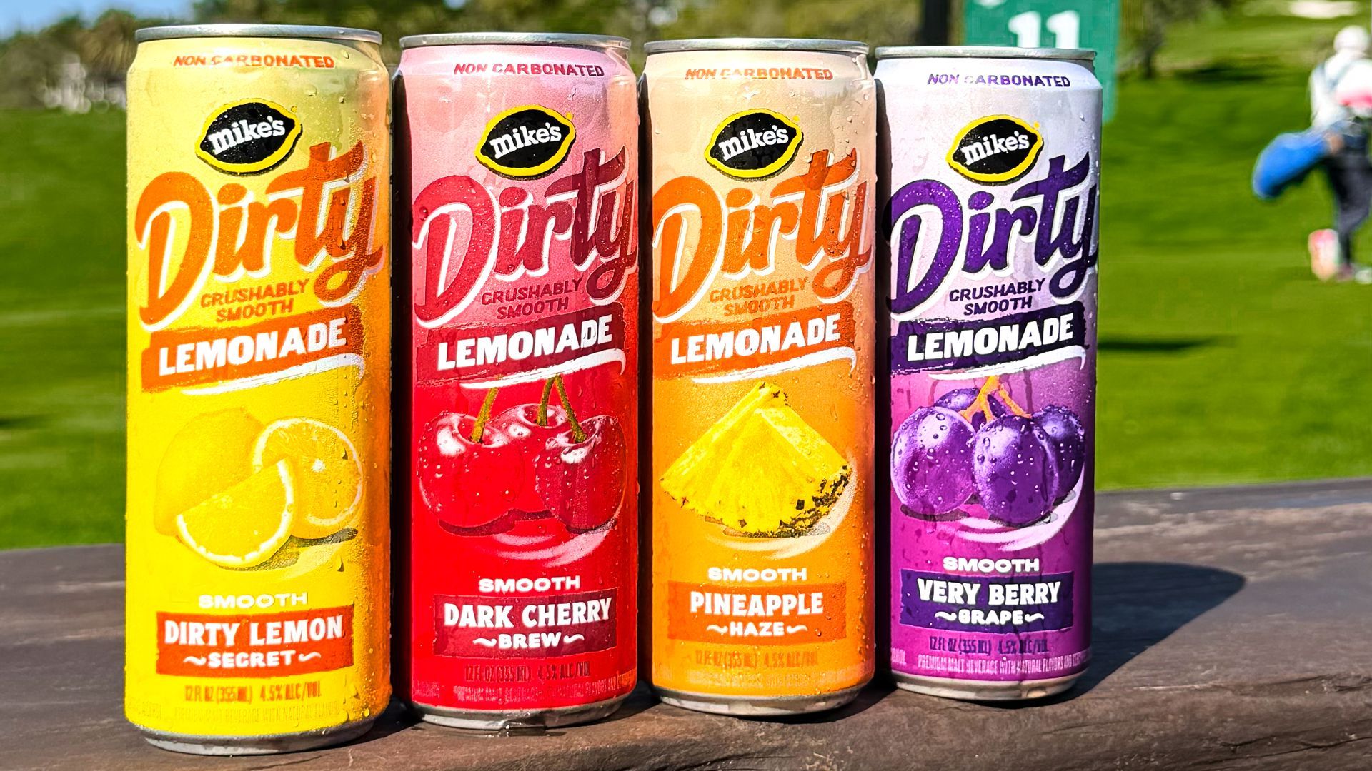 Four colorful cans of Mike's Dirty Lemonade lined up on a wooden table outdoors; left to right: yellow Lemonade, red Cherry, orange Pineapple, purple Berry flavors, with a grassy park in the background.