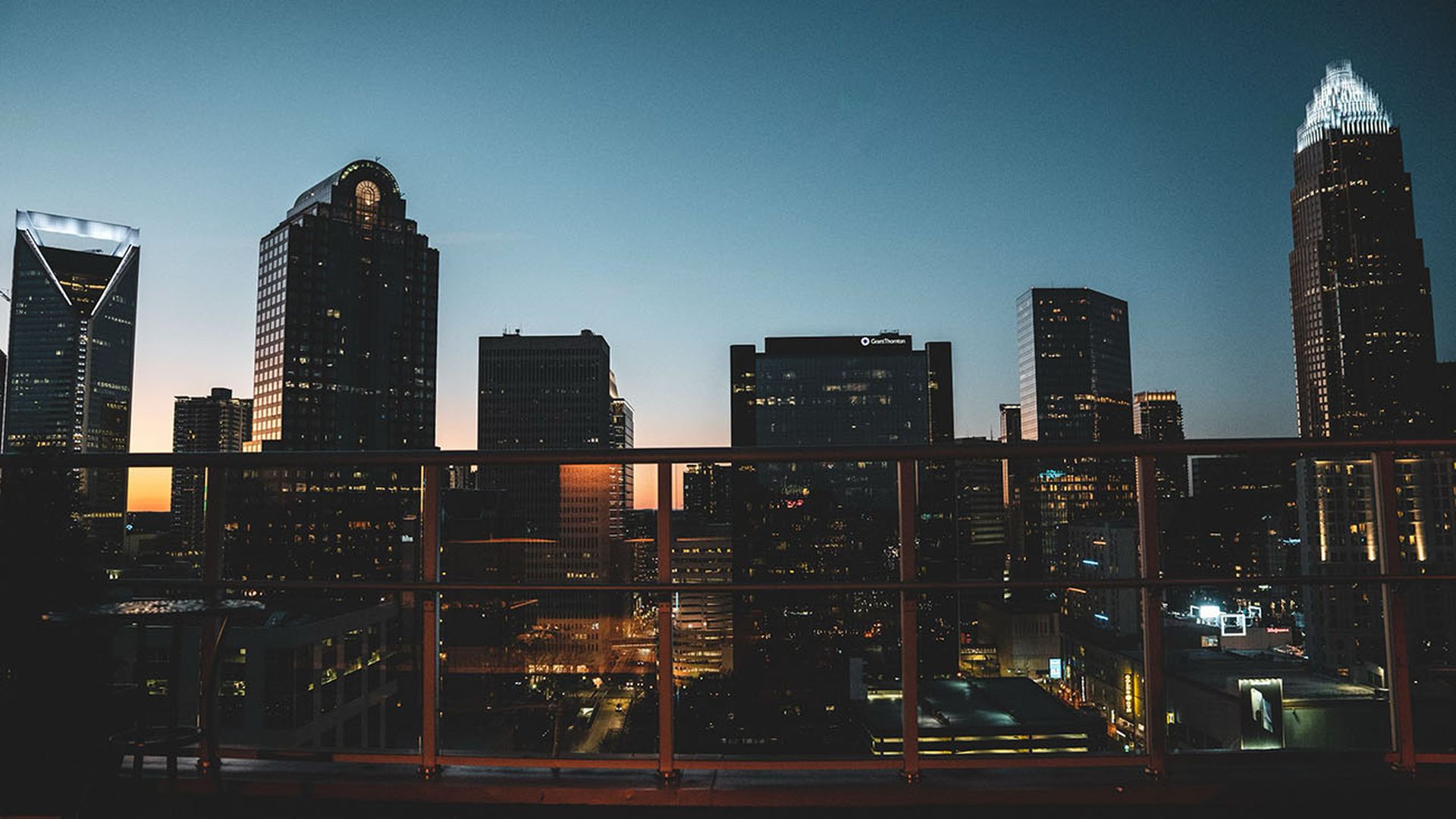 Charlotte City skyline at dusk with illuminated skyscrapers and a clear sky, viewed from behind a glass railing on a rooftop or balcony.