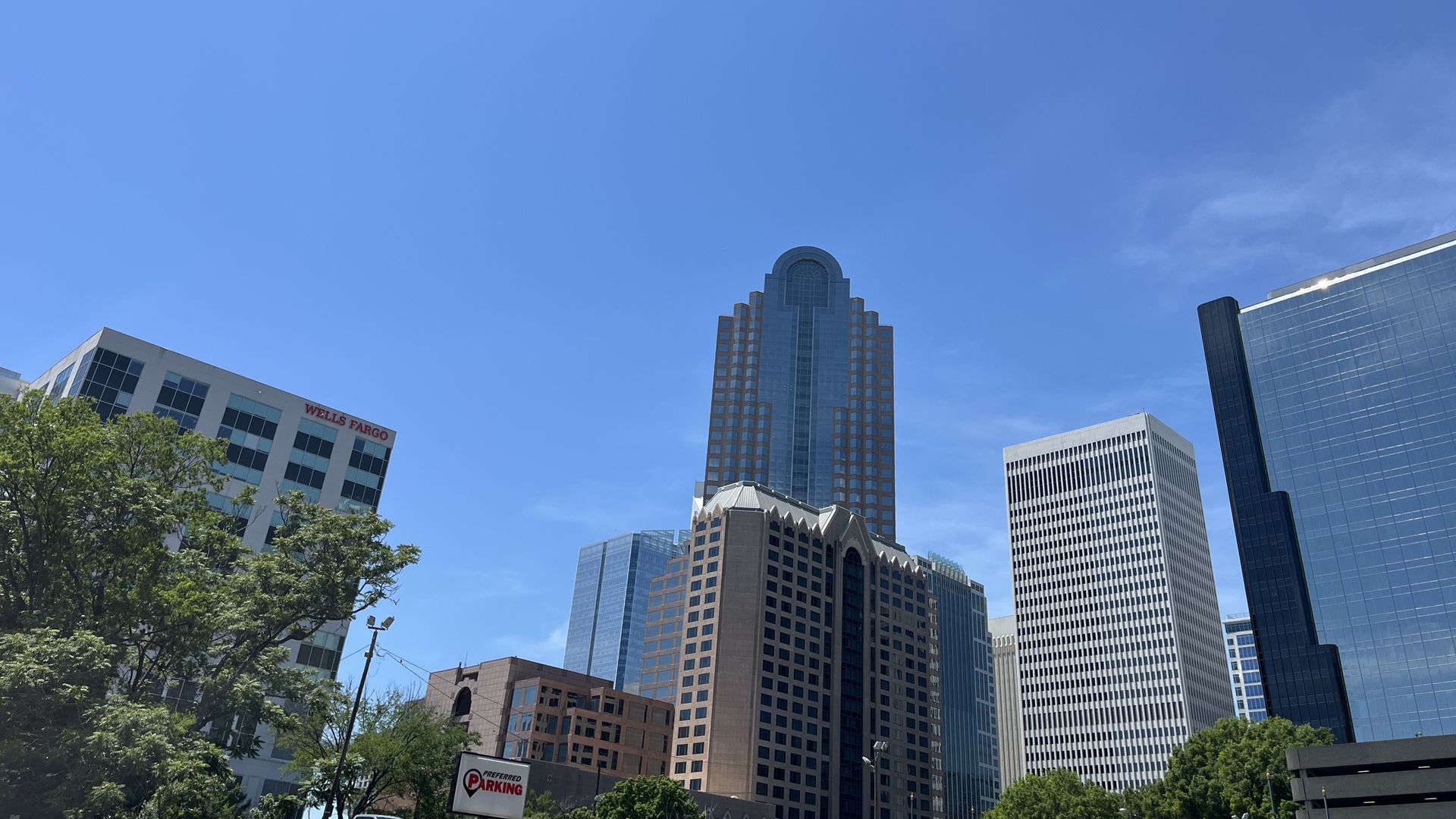 Clear blue sky above a city skyline of tall glass-and-stone skyscrapers, including a central tower with an arched top. Foreground trees and a Wells Fargo sign on a low-rise building.