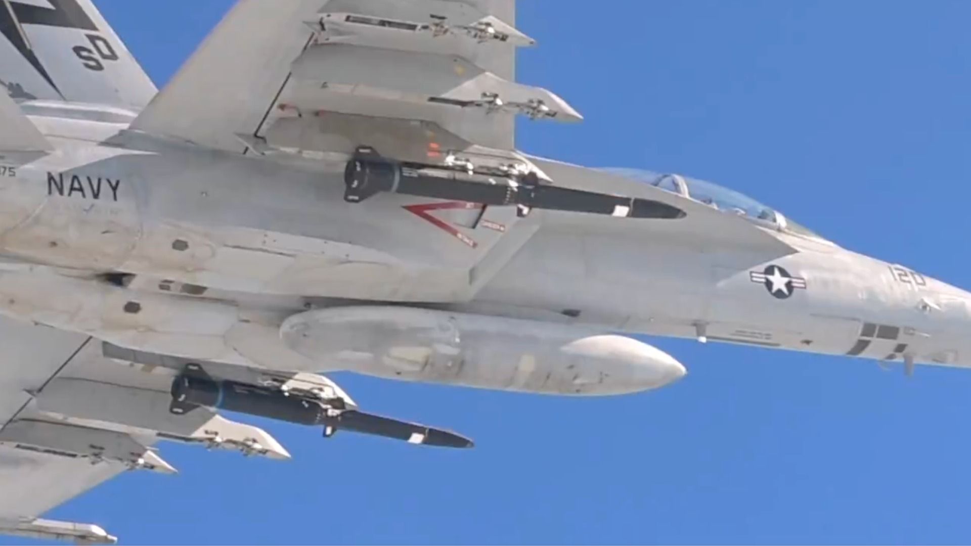 Side view of a gray US Navy fighter aircraft in flight, carrying missiles on underwing pylons.
