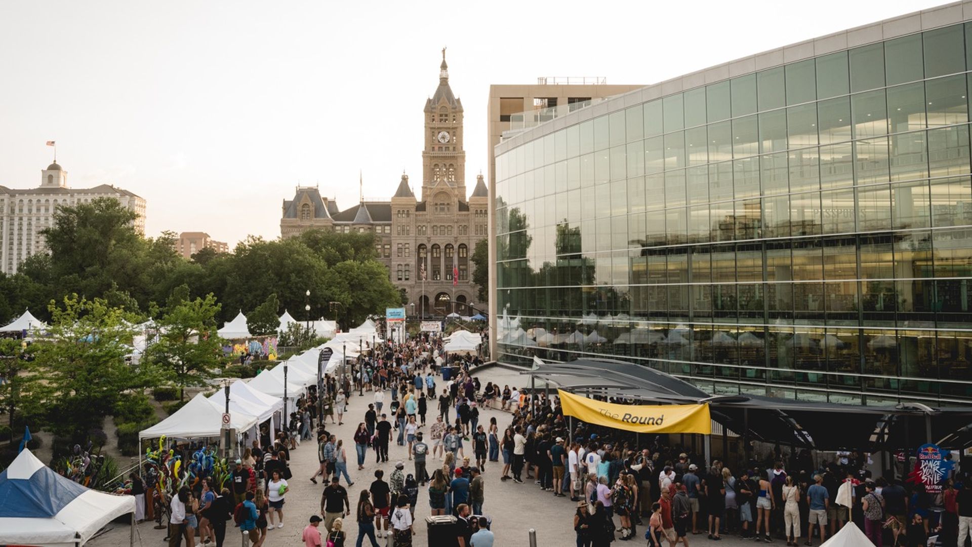 Sunlit city plaza with a glass-fronted modern building on the right, a historic clock-tower in the background, and a busy market-lined walkway filled with white tents and crowds.