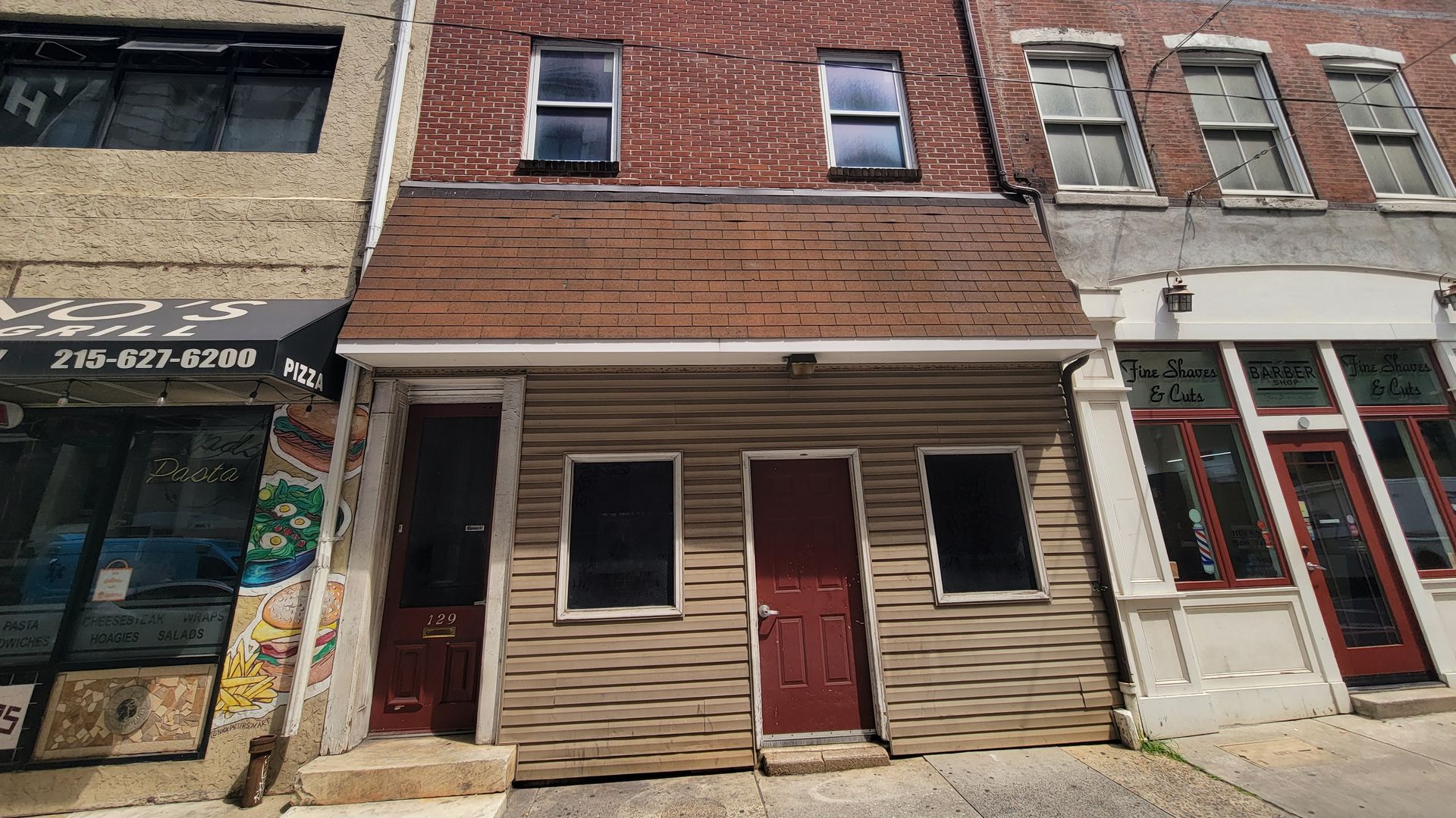 Three adjacent buildings on a city street: a tan siding building with brown doors and two windows, a brick building with four windows, and a white storefront with signs for barber services.