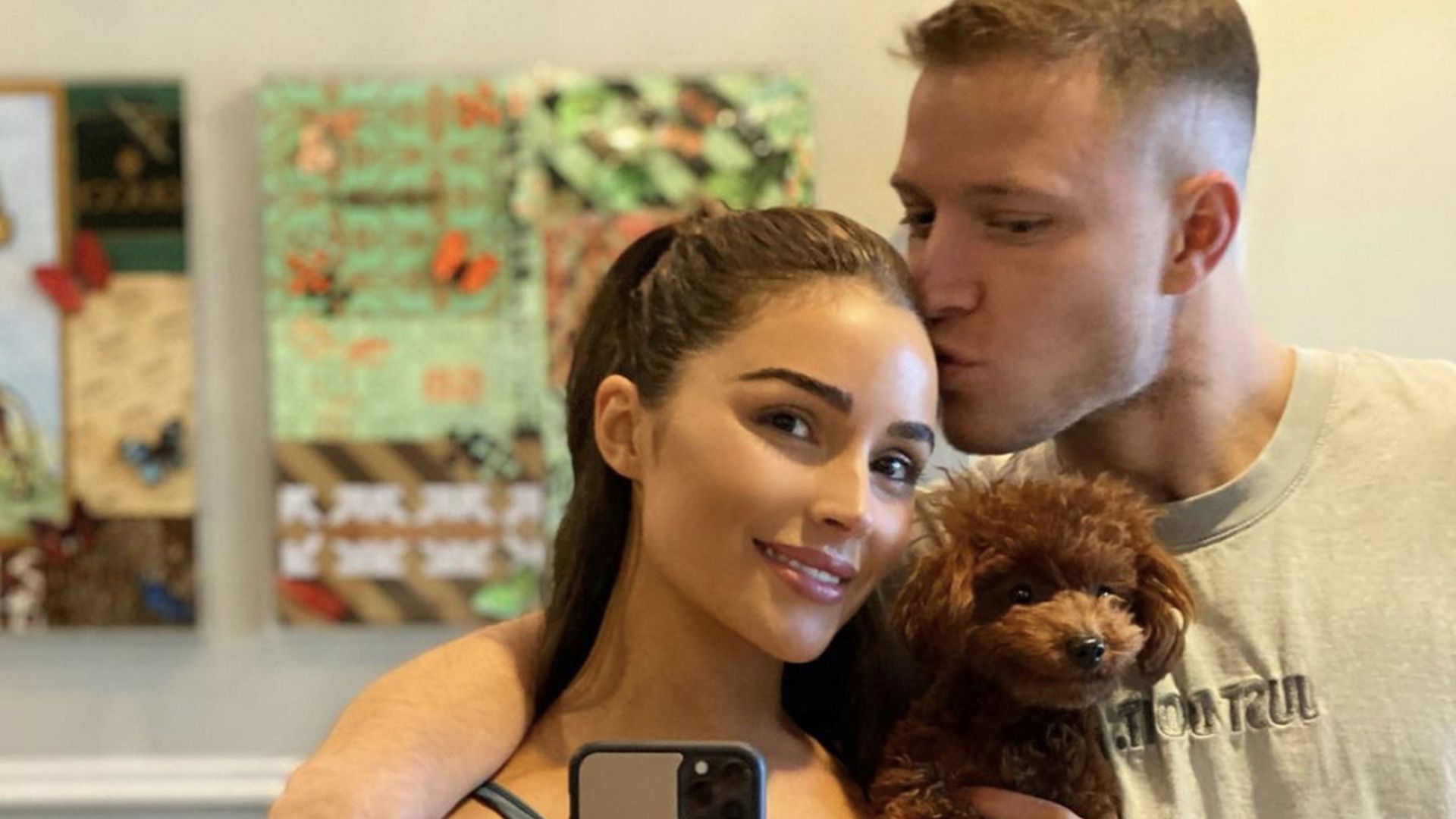 Supermodel Olivia Culpo is holding a phone in her right hand and her toy golden doodle in her left, while being kissed on the forehead by boyfriend Christian McCaffrey