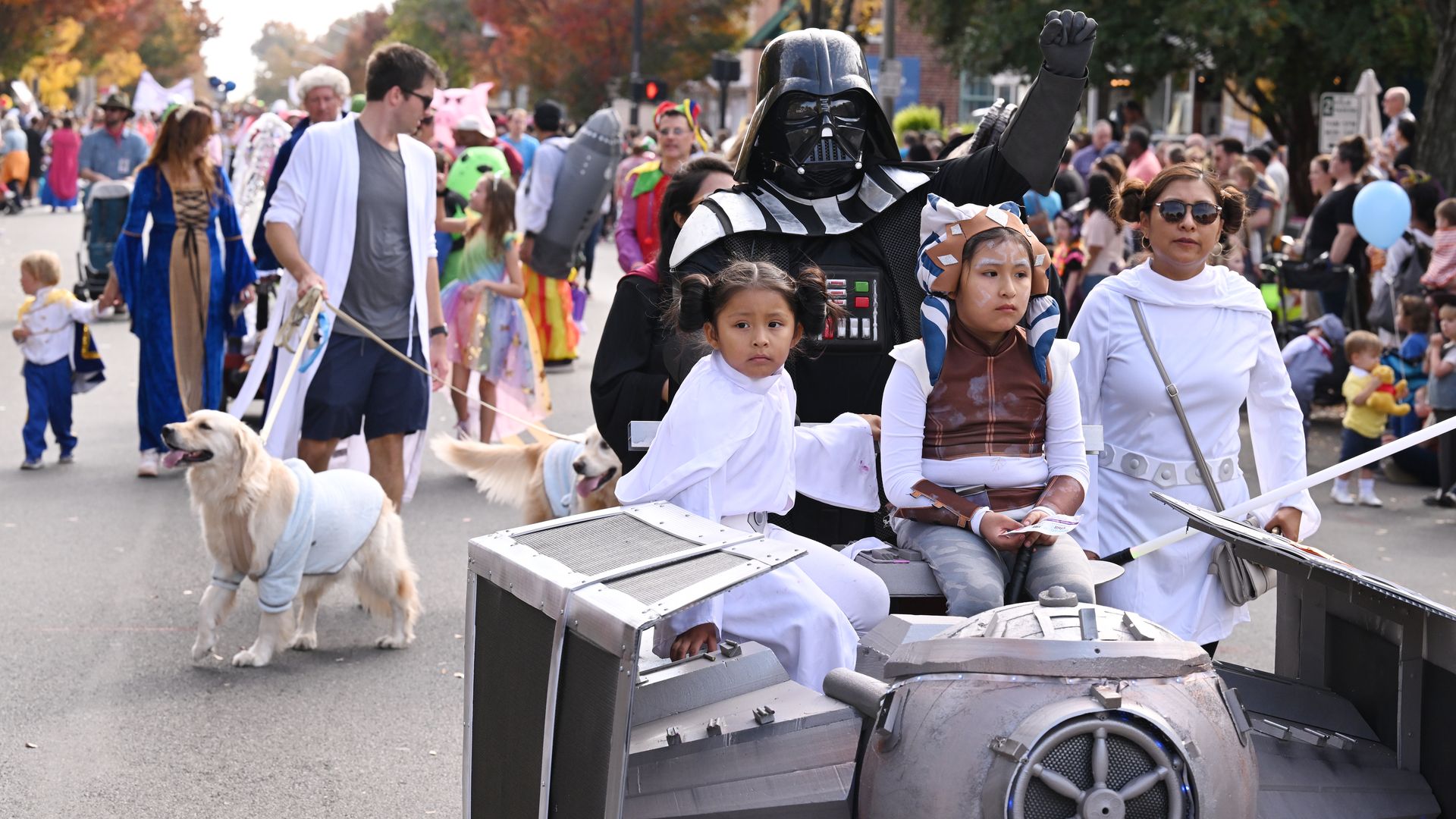Darth Vader in black armor rides a parade float beside two girls in white costumes; a golden retriever in a blue sweater walks ahead as costumed spectators line a tree-lined autumn street.