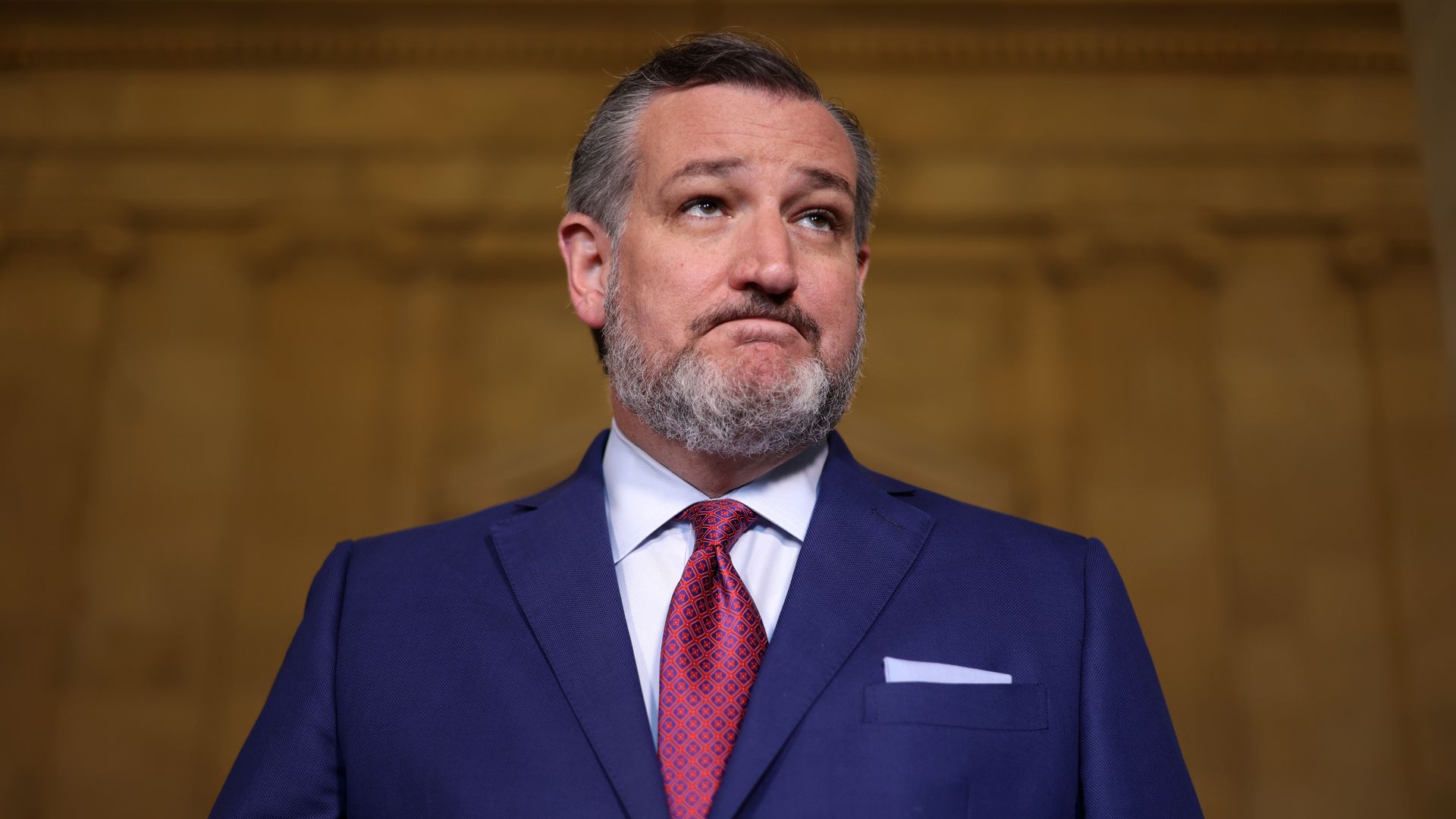 A man with a light beard, in a blue suit and maroon tie, looks somewhat annoyed, in an official setting. 