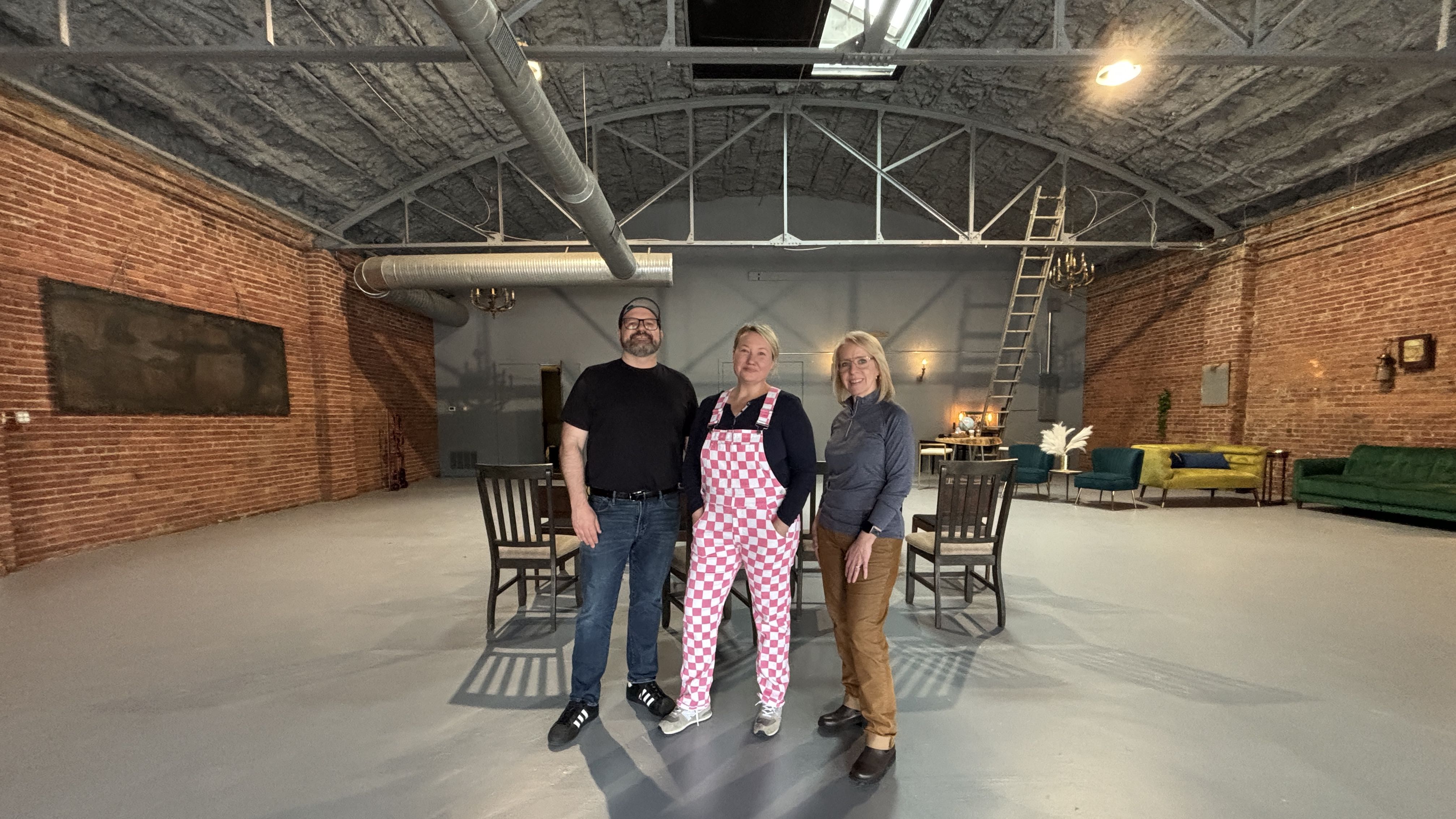 Three people stand in a large brick-walled studio with exposed ducts. A man in black, a woman in pink-checkered overalls, and a woman in gray jacket pose near chairs and a ladder in back.