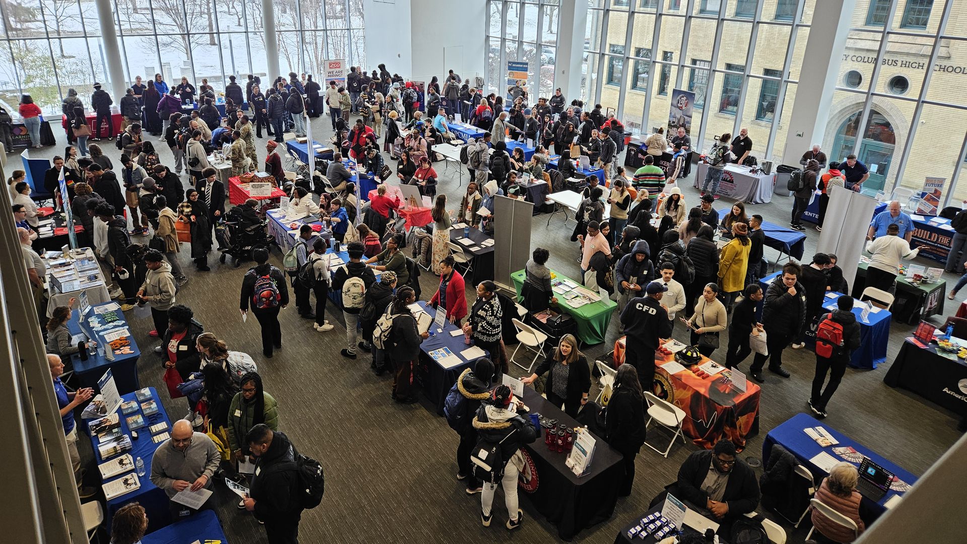 Busy job fair inside a large room with floor-to-ceiling windows and many people interacting with representatives at tables covered with colorful cloths.