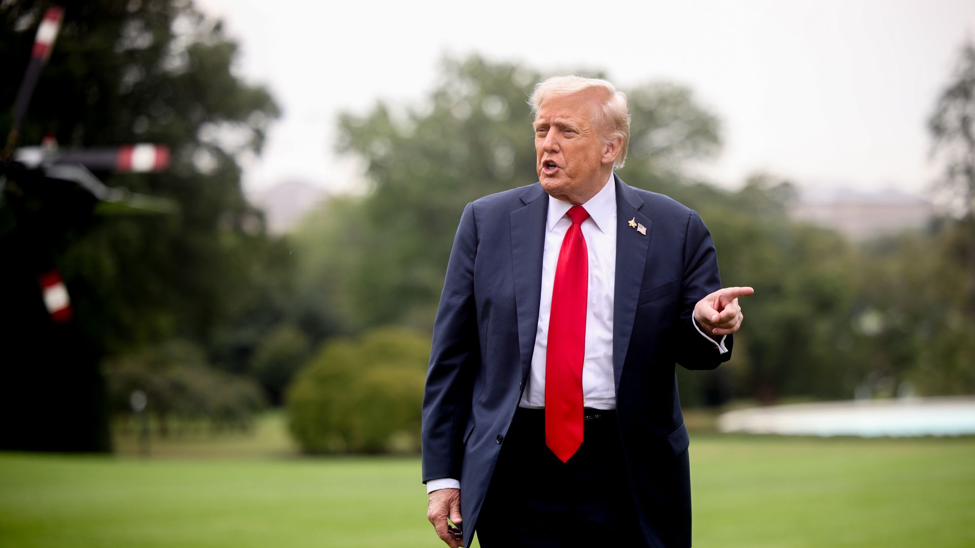 Trump points as he walks across the White House lawn wearing a dark suit and a red tie.