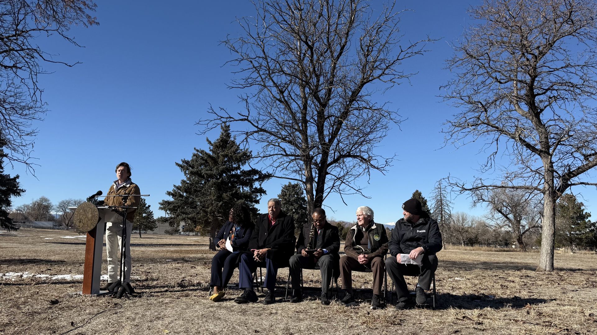 A man speaks at lectern at a park while other people are seated nearby. 