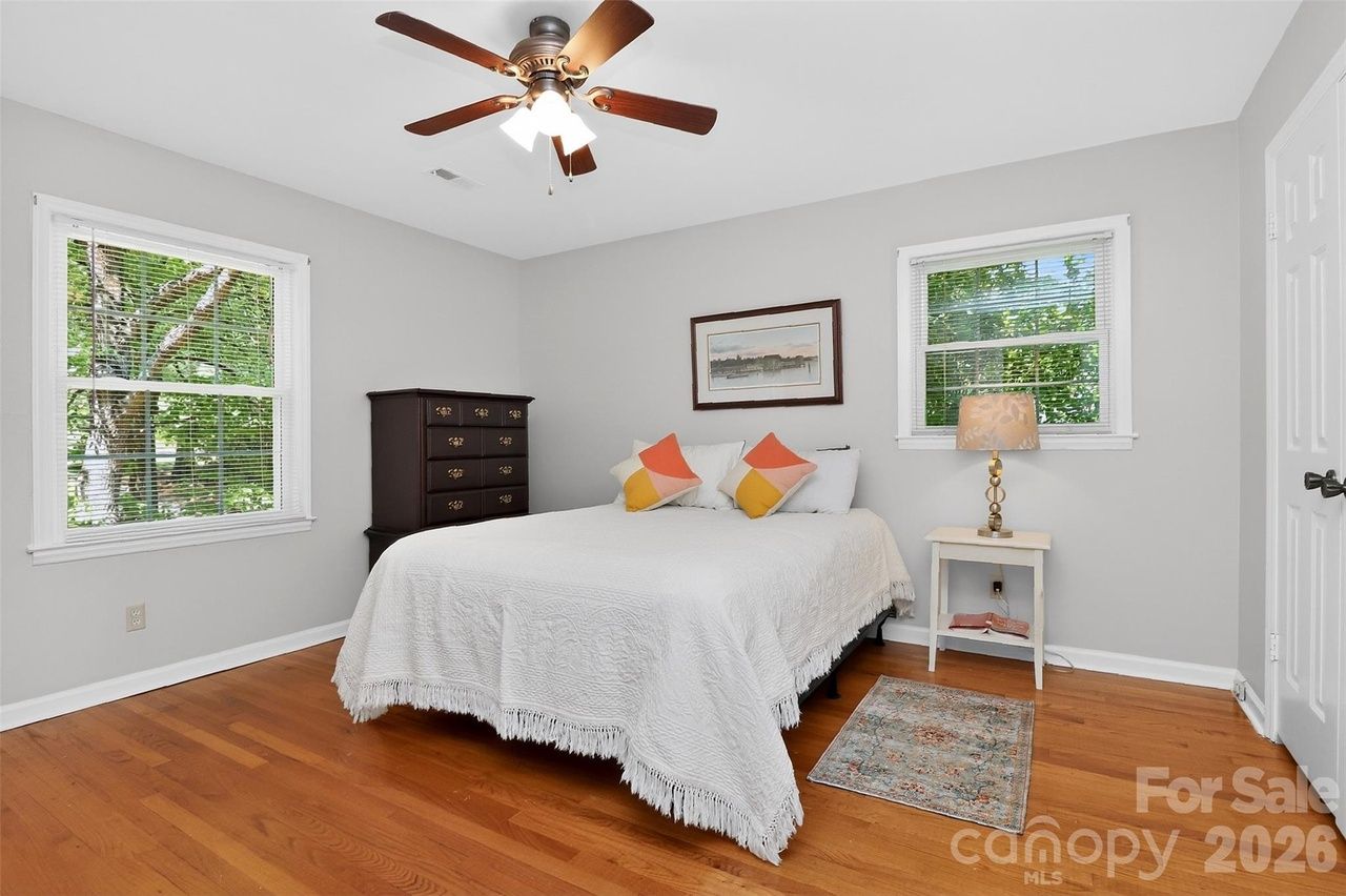Bright bedroom with gray walls, white bedspread and fringe. Dark wood dresser left, white nightstand with lamp right. Two windows, ceiling fan, hardwood floors, small rug.