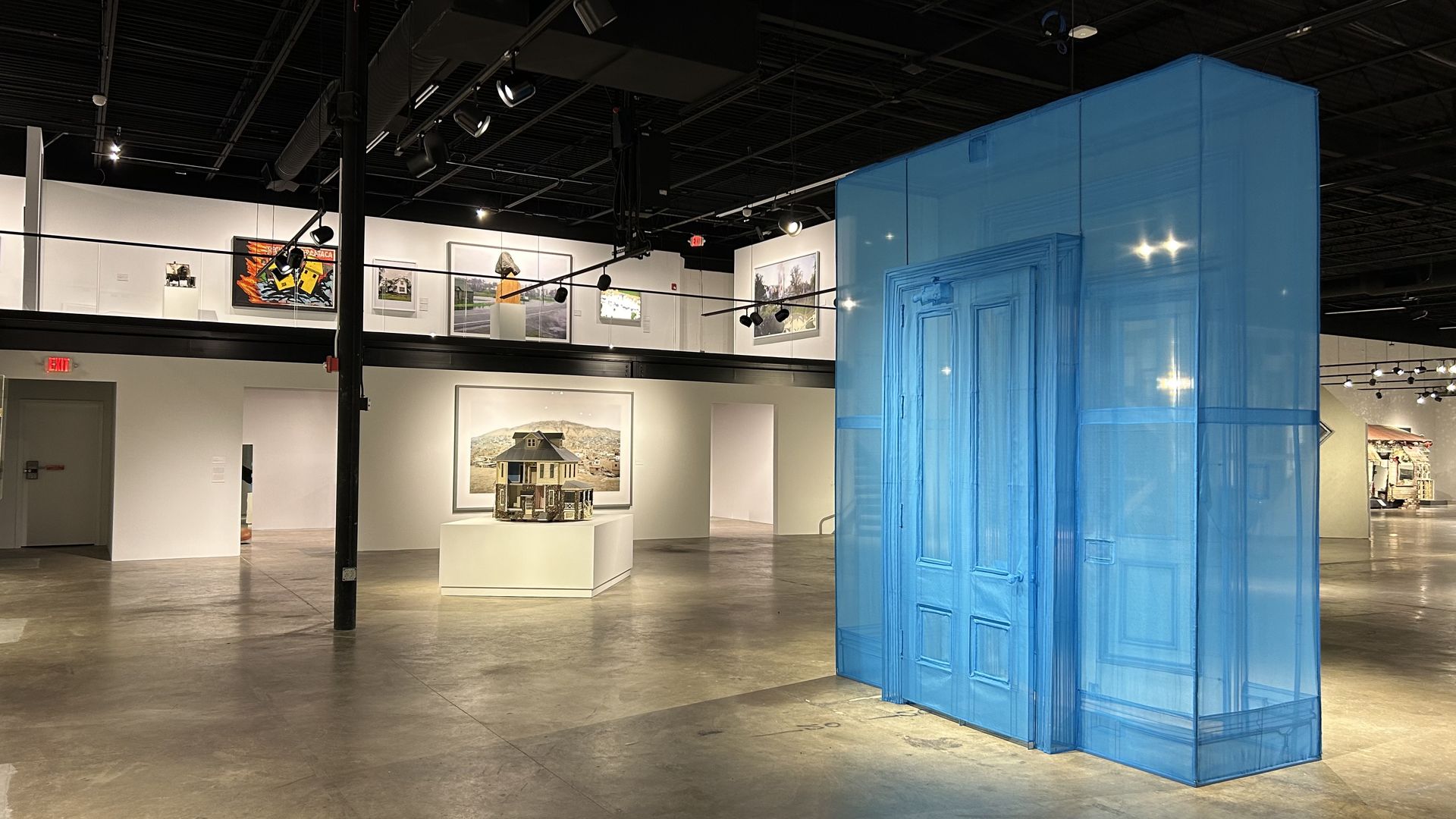 A blue translucent fabric art work of a detailed entranceway is prominently displayed in the center of a white wall art gallery