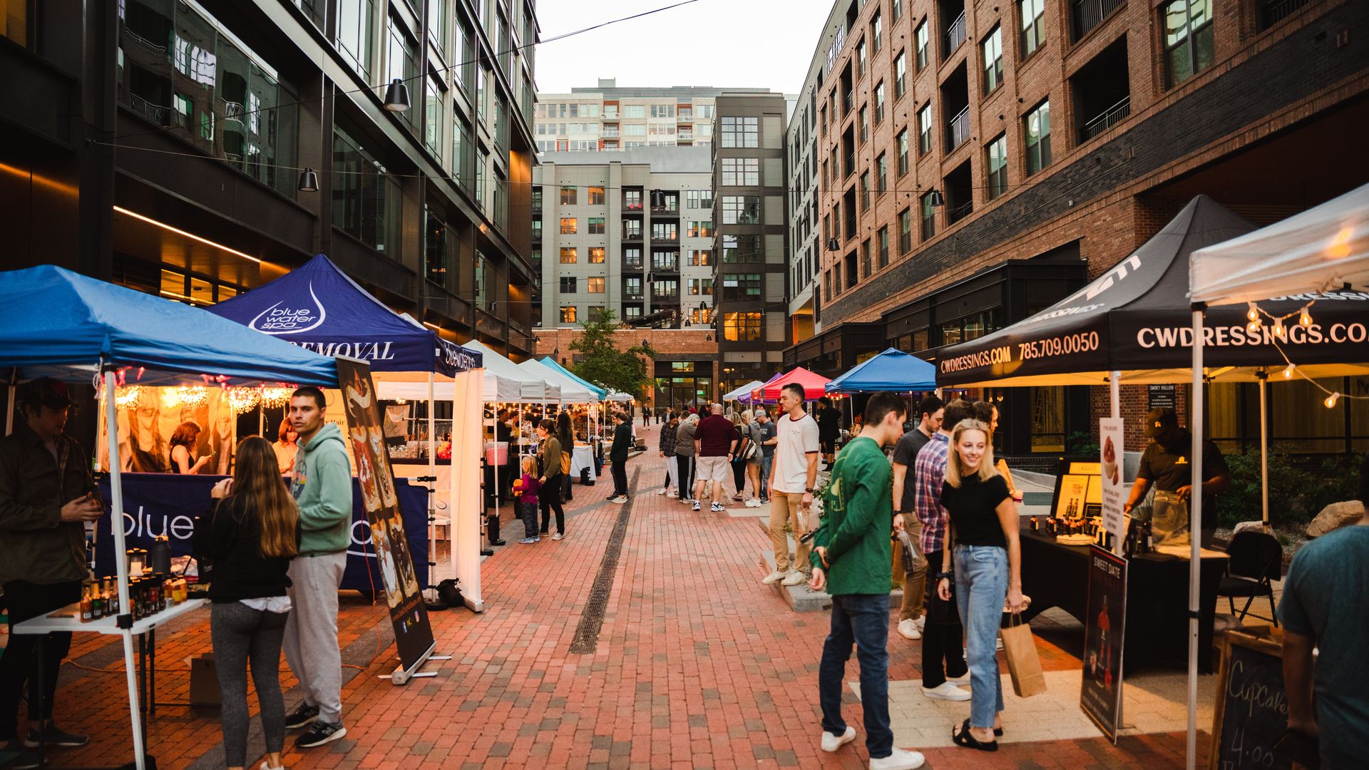 People browsing stalls at an outdoor market between tall modern buildings during dusk, with colorful tents and warm lights creating a lively urban atmosphere.