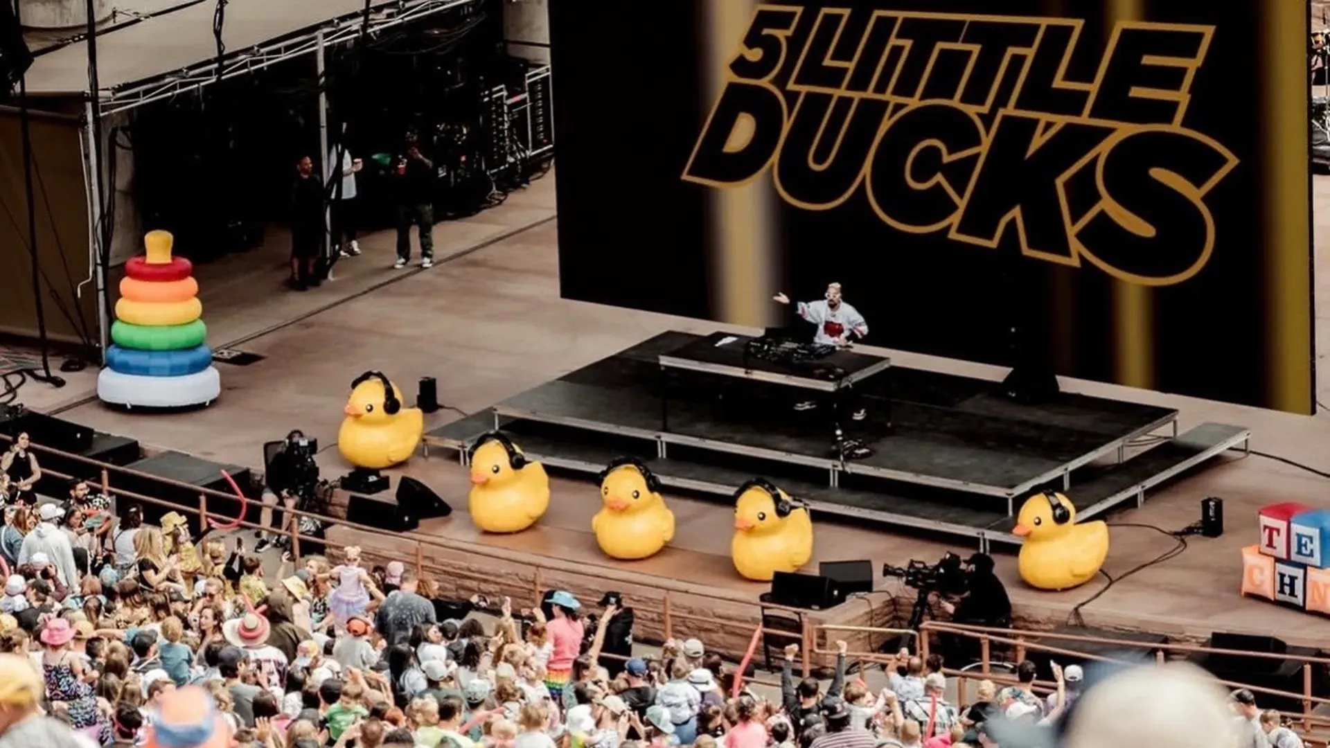 DJ performs on a raised stage with a bold orange backdrop; yellow inflatable ducks wearing headphones line the front, and a tall rainbow ring stack sits to the left, as a large crowd watches.