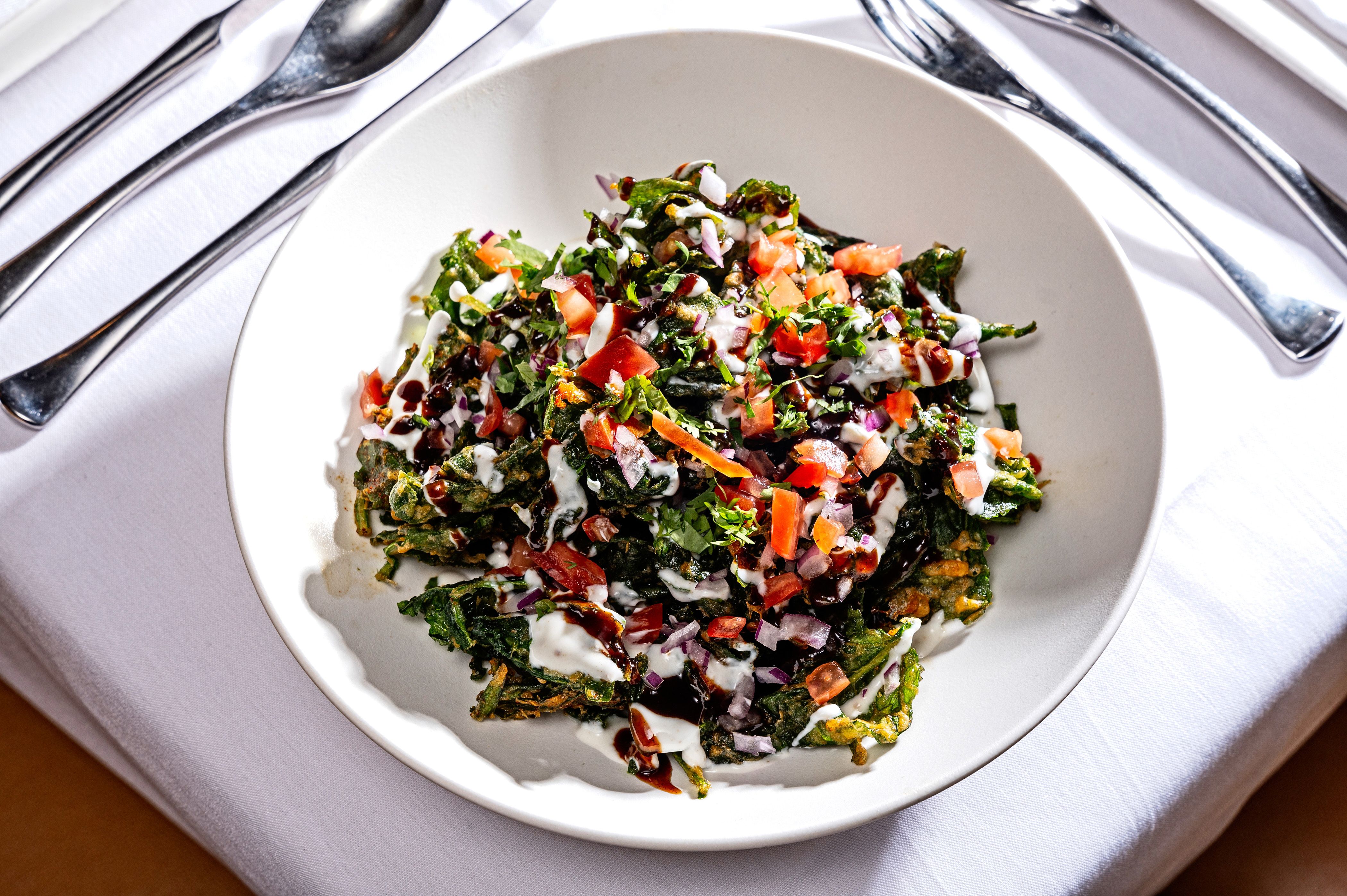 White bowl filled with crispy green spinach pakoras topped with diced tomatoes, chopped onions, cilantro, and drizzled with white and dark brown sauces on a white tablecloth next to silver cutlery.