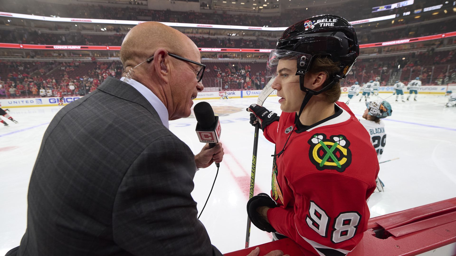 Photo of a man in a suit interviewing a hockey player on the ice.