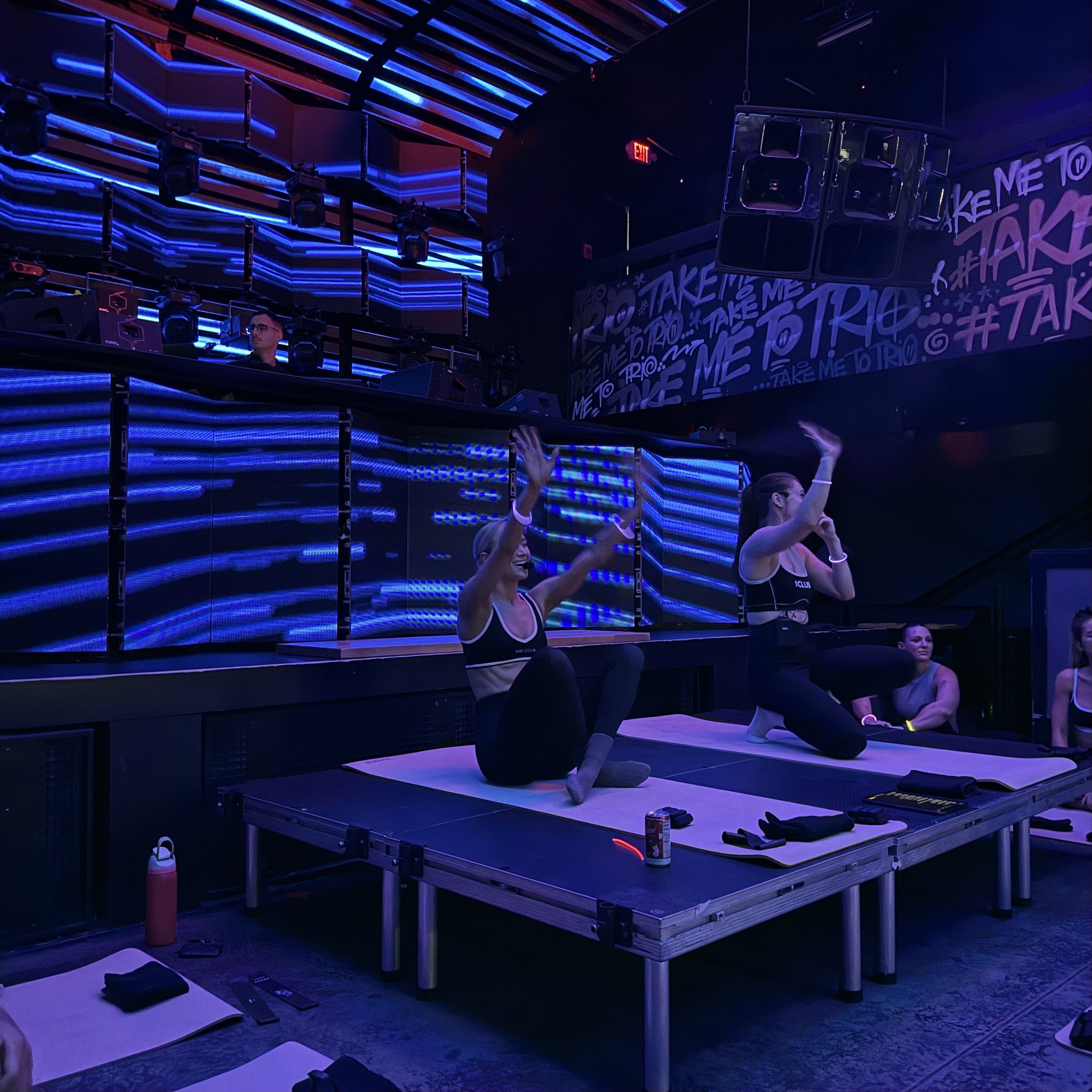 Two women in black athletic wear exercise on mats on a stage with blue and purple neon lights and graffiti reading "Take me to Trio" behind them, with a DJ in the background.