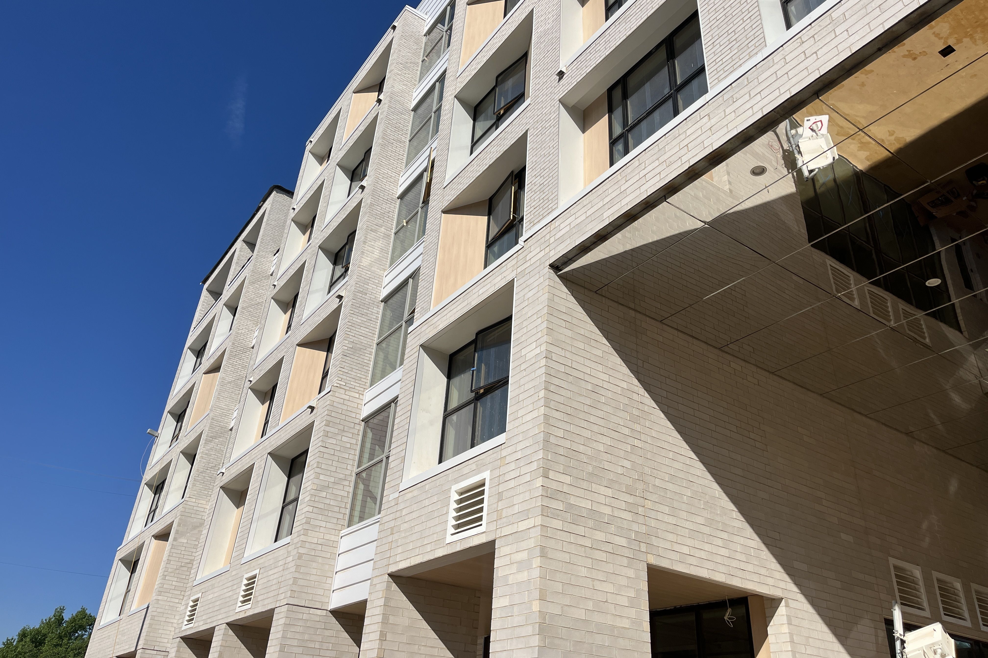 Low-angle view of a modern beige brick apartment building with square windows and recessed balconies, under a clear blue sky with shadows across the entrance area.