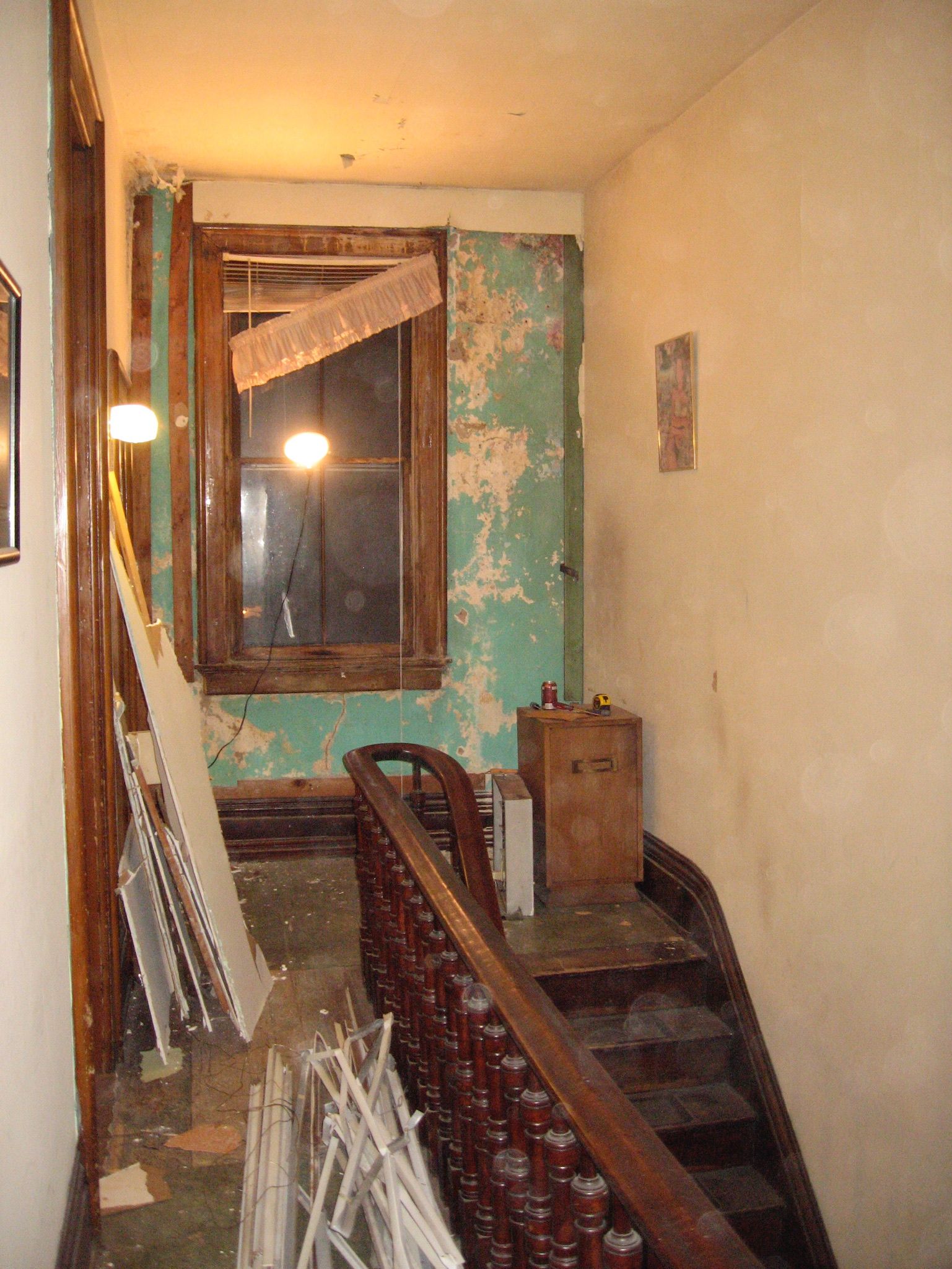A narrow, dim hallway in an old house under renovation. Peeling teal wallpaper, a curved wooden banister with red spindles, a window with blinds, and scattered debris with a small cabinet.
