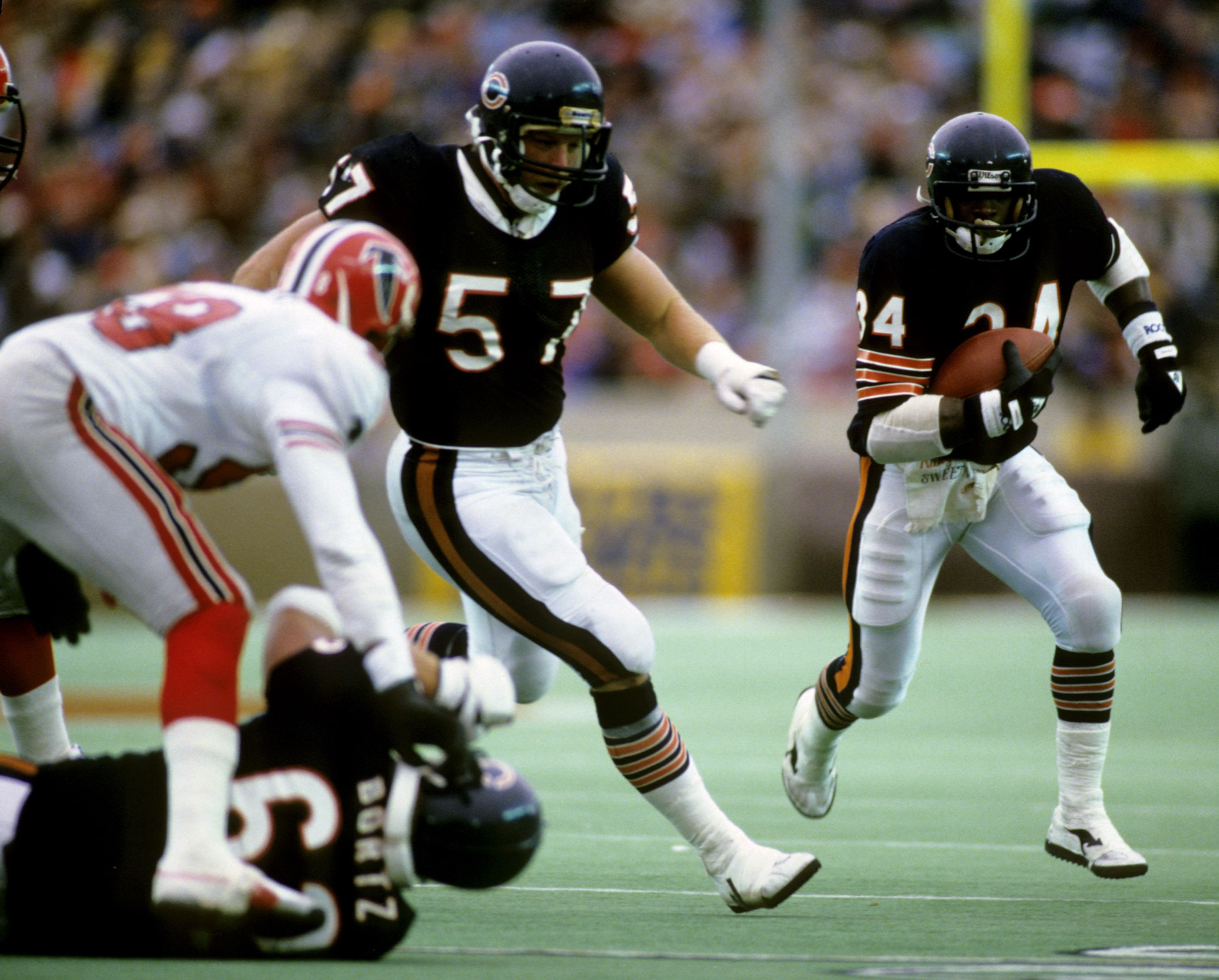 Chicago Bears player #34 runs with football while teammate #57 blocks a defender in white and red uniform during a game on green field.