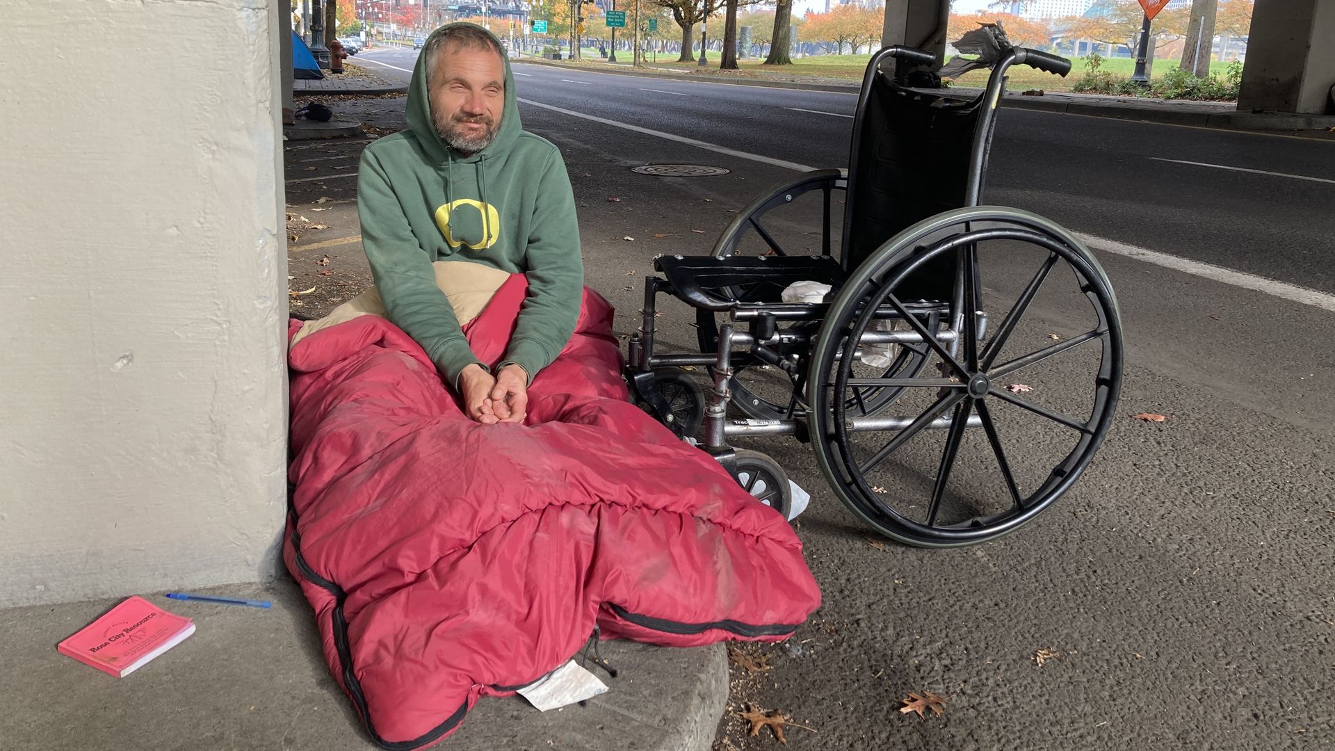 A man in a green hoodie with a red sleeping bag over his lap sits on the sidewalk next to his wheelchair.