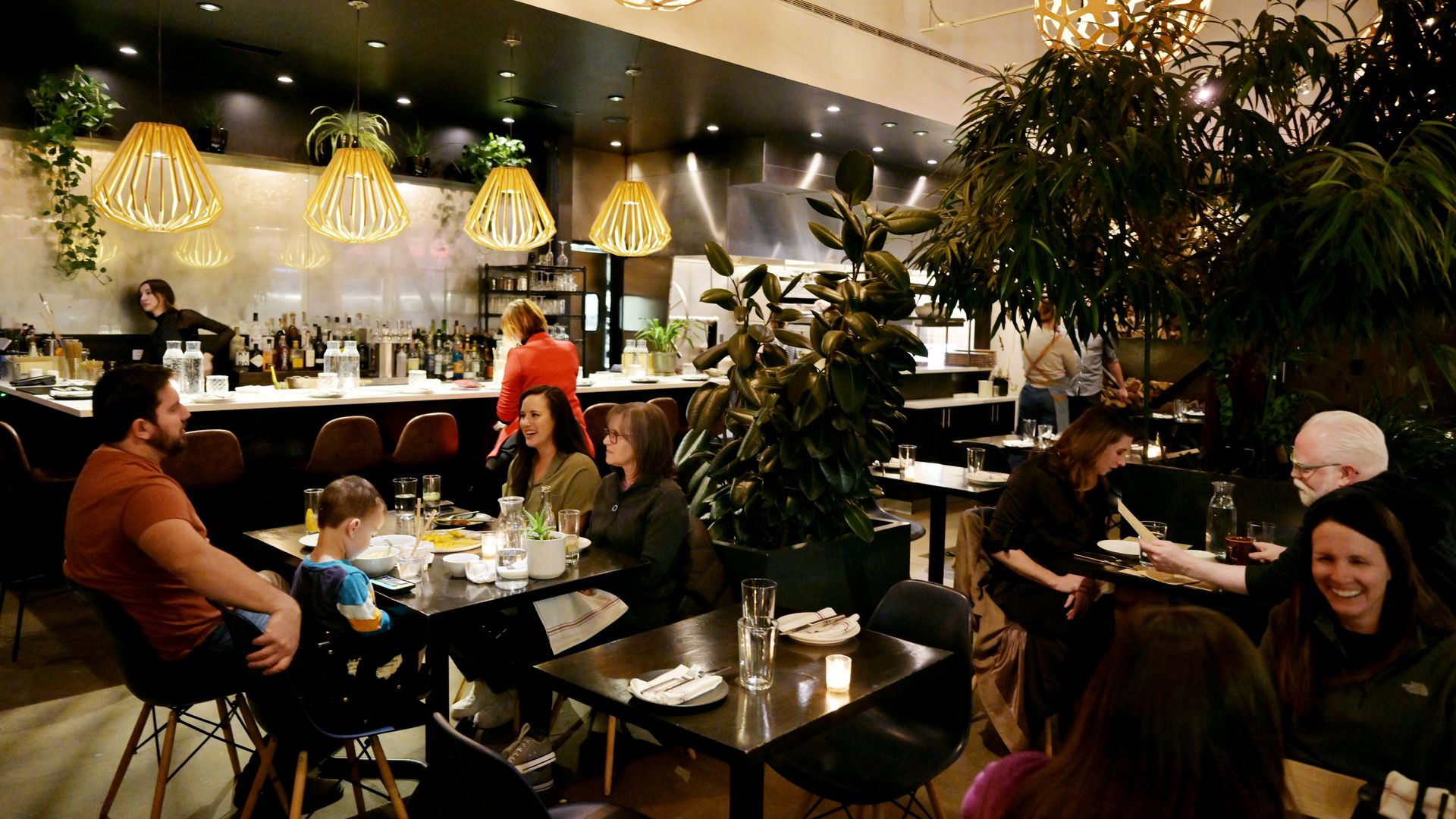 Cozy restaurant with warm yellow lights, multiple diners including a family with a child, green plants, and a bar area with bottles and staff in the background.