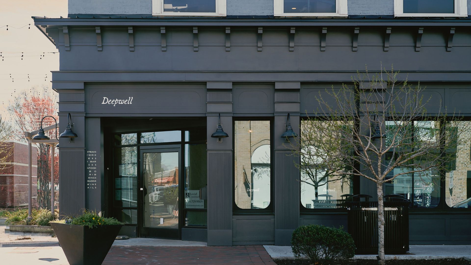 A dark gray brick storefront with decorative cornice and the word Deepwell on the facade; glass doors, planters, and leafless trees outside; string lights hang overhead.