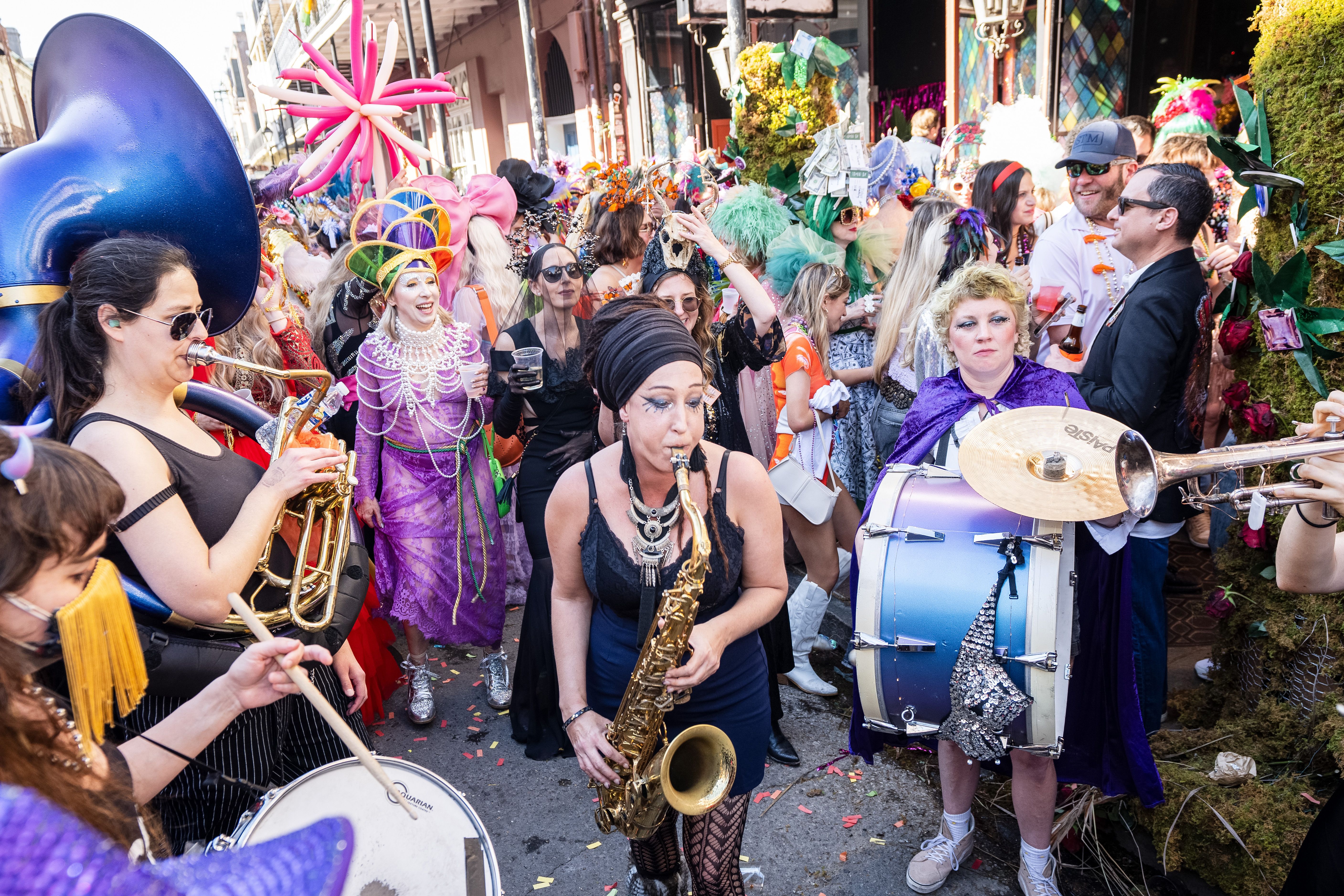 Colorful street parade with musicians playing saxophone, tuba, trumpet, and drums. People in vibrant costumes, including purple, black, and green outfits, some with elaborate headpieces.