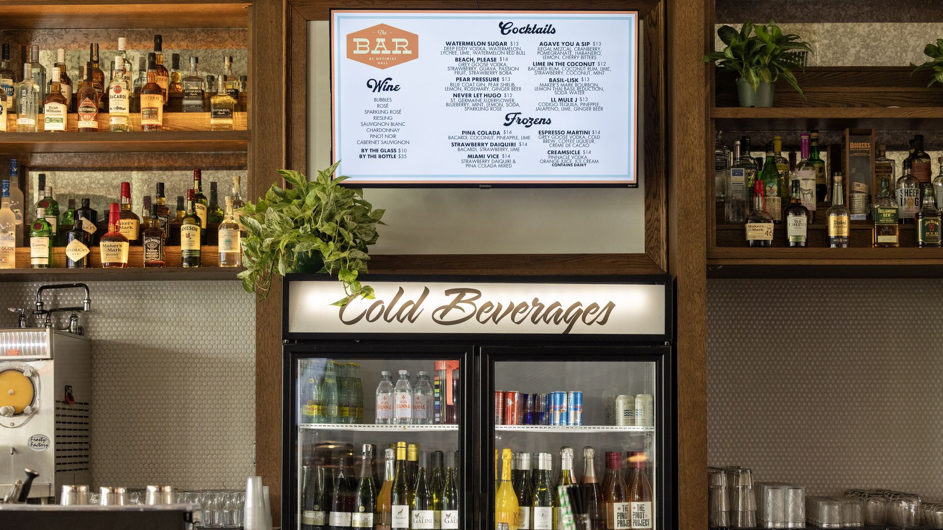 Bar area with wooden shelves holding various liquor bottles, a menu board displaying cocktails and wines, a cold beverages fridge with assorted drinks, and plants on top.