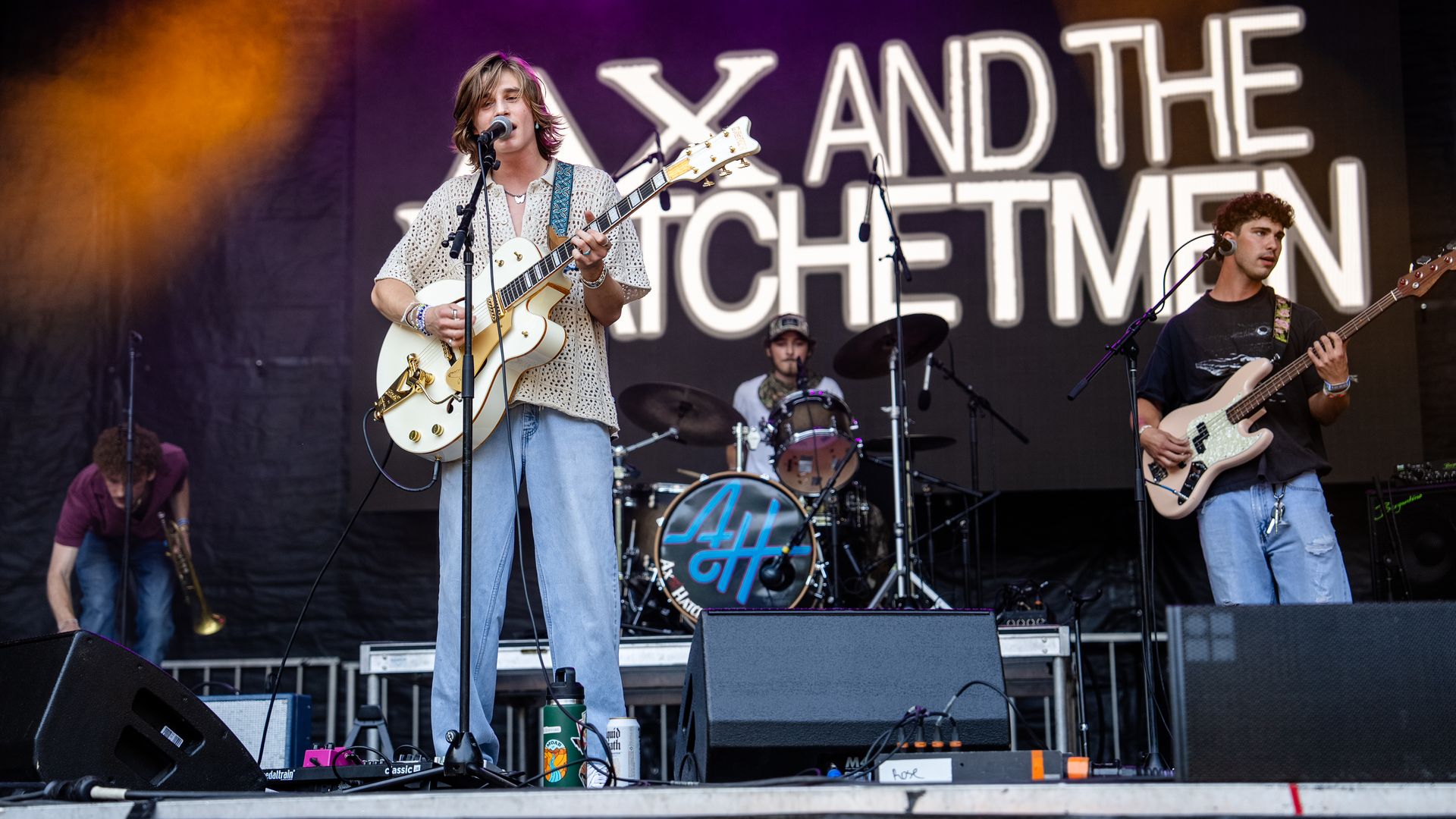 Outdoor band on stage; frontman with white guitar and patterned shirt, brown hair, singing into mic. Purple/orange lights, large backdrop text; bassist on right, drummer behind.