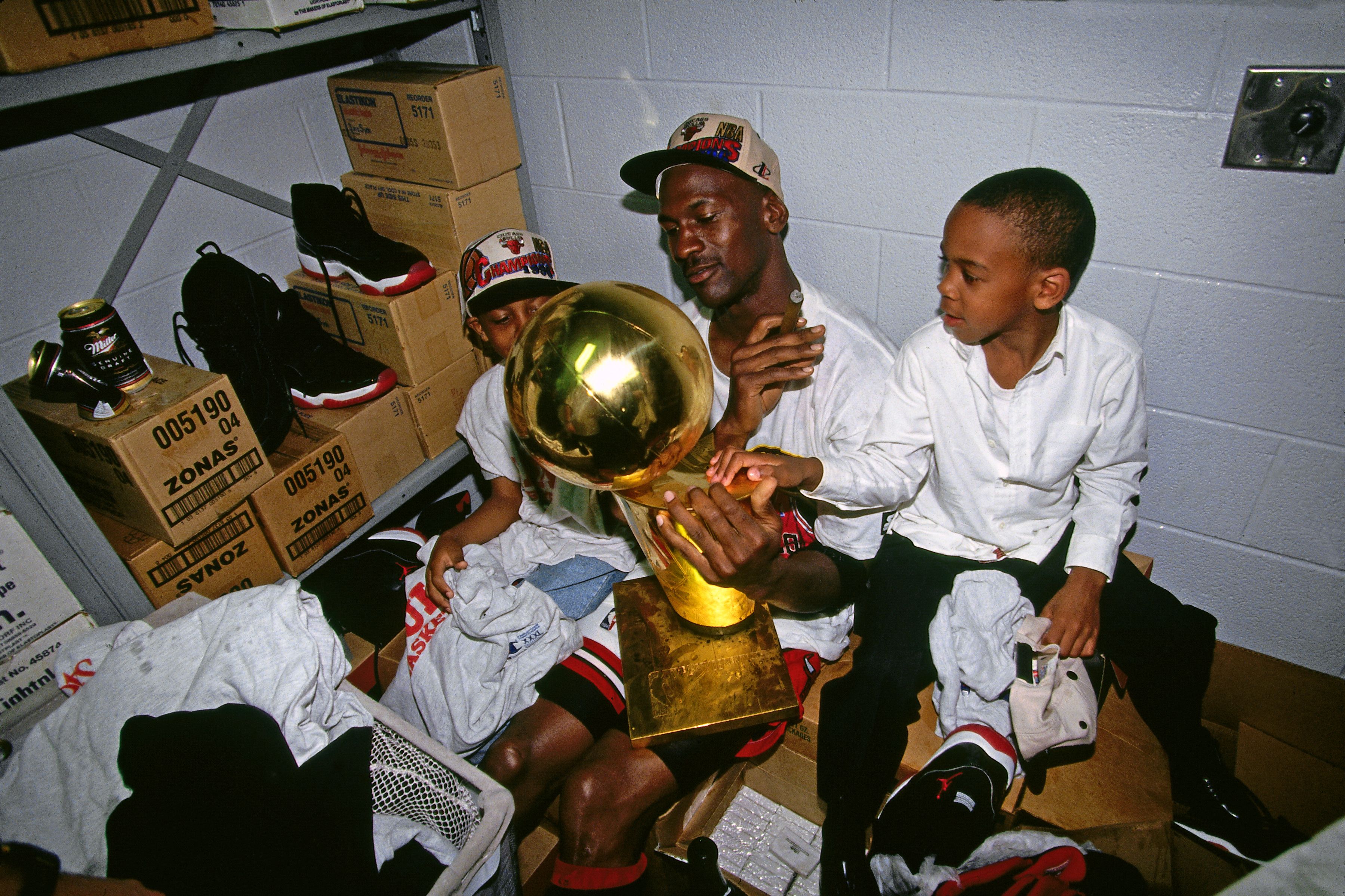 People in a storage room celebrate with a large, shiny gold trophy; a man in a white shirt and cap holds it while a boy looks on, surrounded by boxes, sneakers, and clothes.