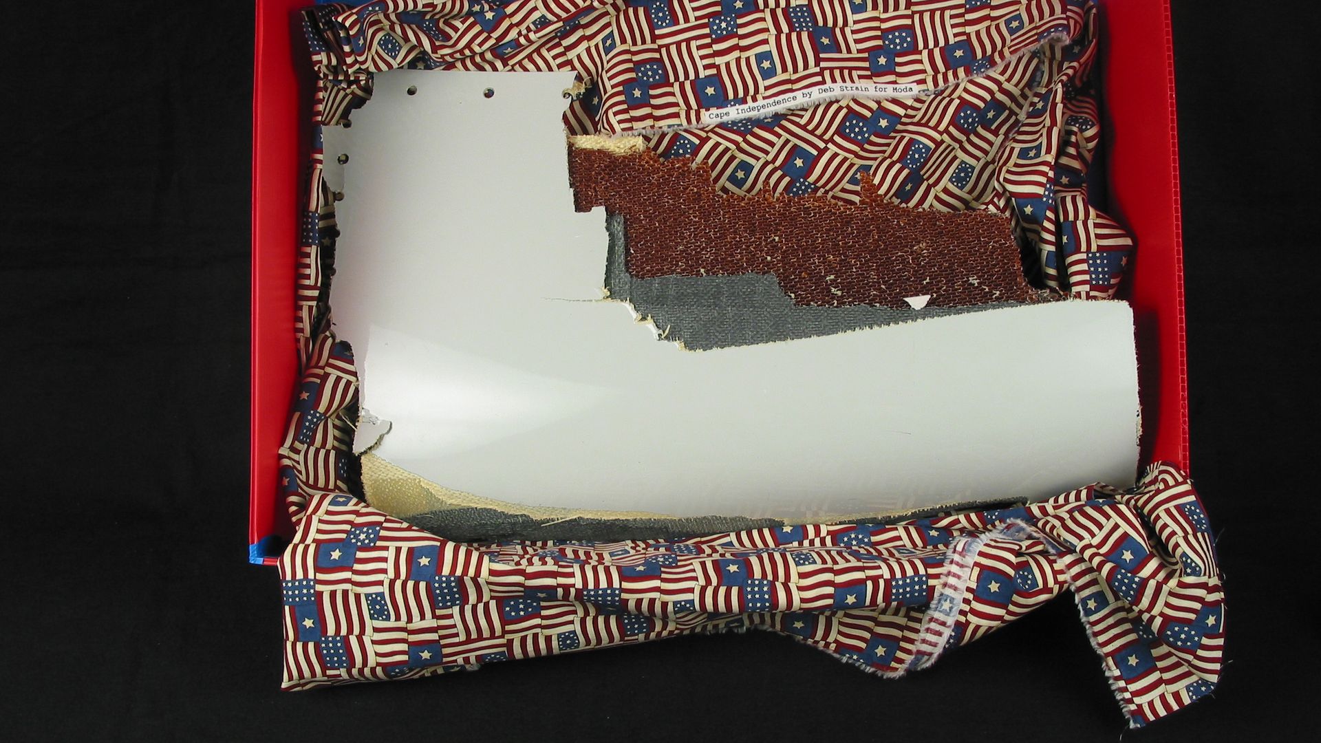 Red box with blue edges sits on a black background. Inside is crumpled patriotic fabric with an American flag pattern, a torn white panel, and a brown textured patch; a small label is visible.