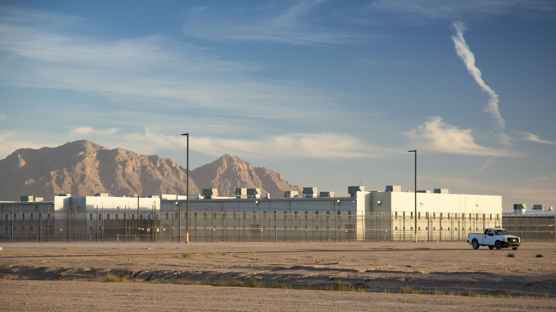A large, low-slung white prison-style building surrounded by wire fencing in a brown desert landscape, with mountains in the background.