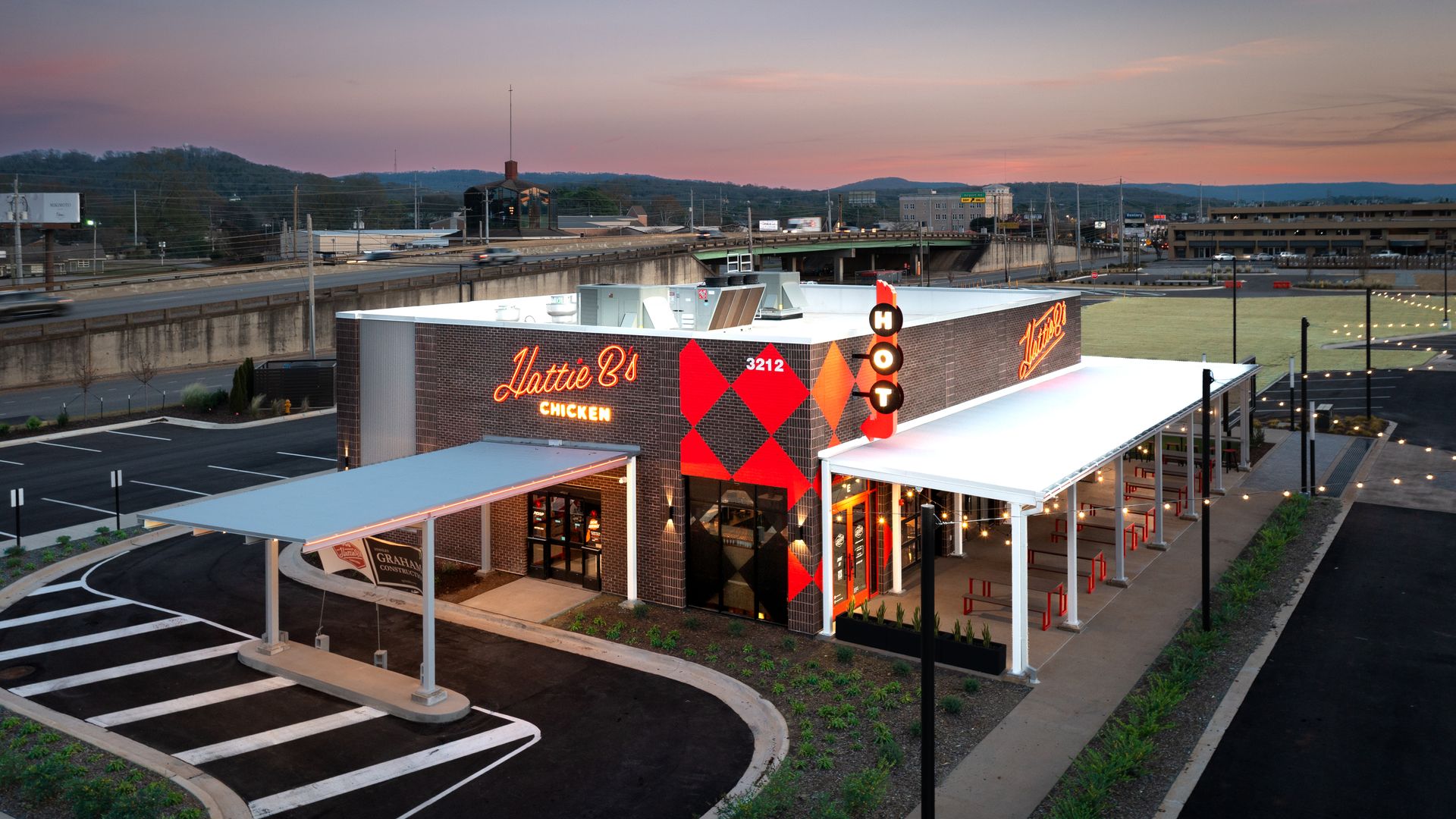 Aerial view of Hattie B's Chicken restaurant at dusk with bright orange neon signage, red and black diamond pattern on the brick building, outdoor covered seating with red stools, and nearby highway.