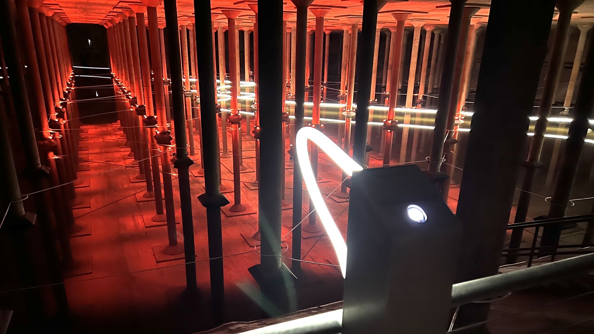 Red-lit room filled with rows of tall vertical pillars forming a tunnel; water reflects the pillars. A bright white neon tube curves across the foreground.
