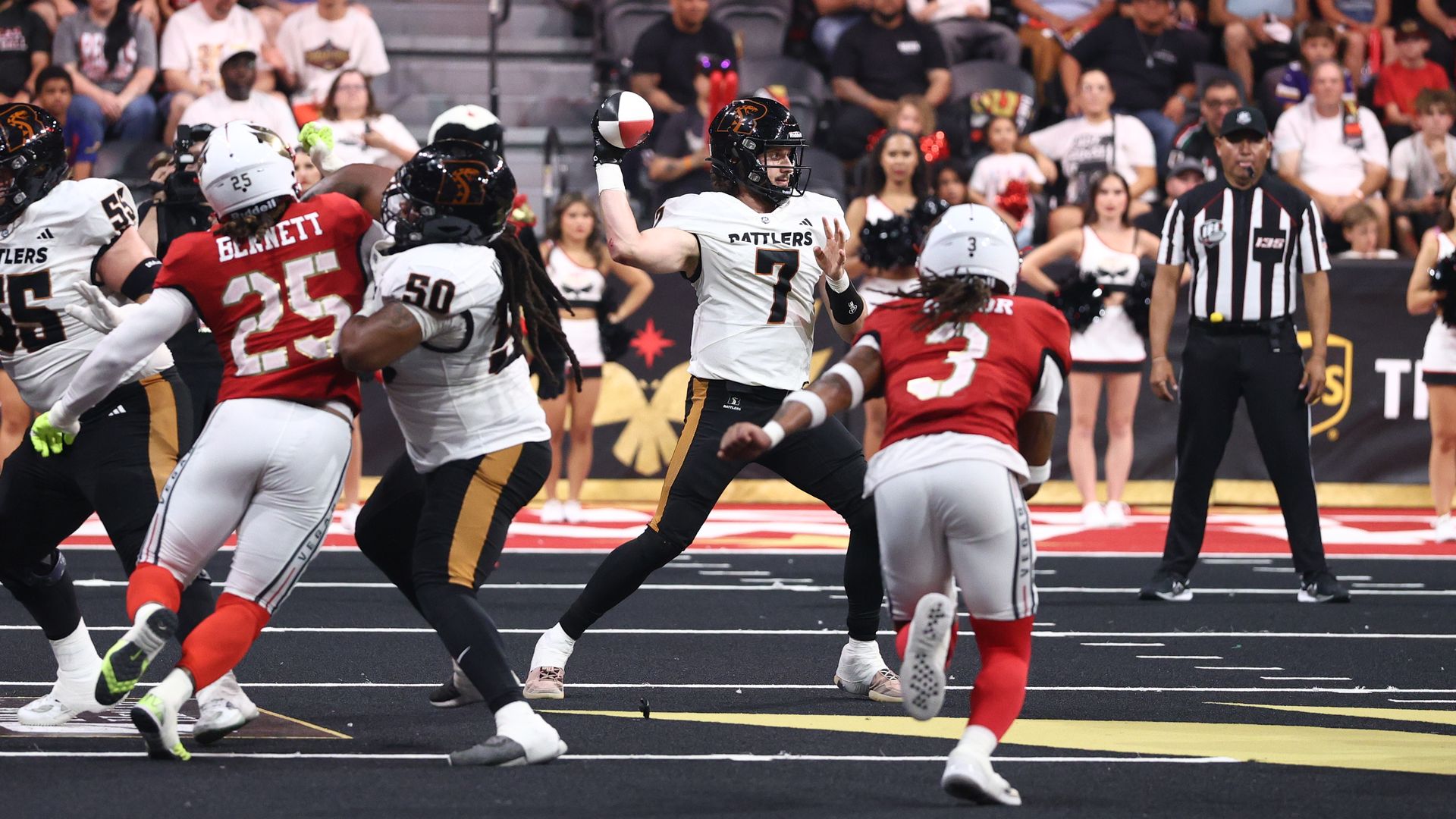 Quarterback in white "Rattlers" jersey (No. 7) standing on a black surface throws a pass as red-jersey defenders converge; crowd in stands, yellow goalpost in background, referee on the right.