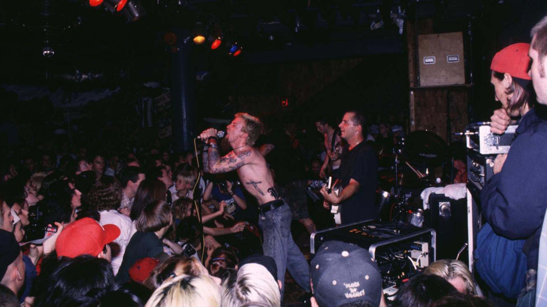 A man on stage with no shirt sings into a microphone in front of a mass of fans