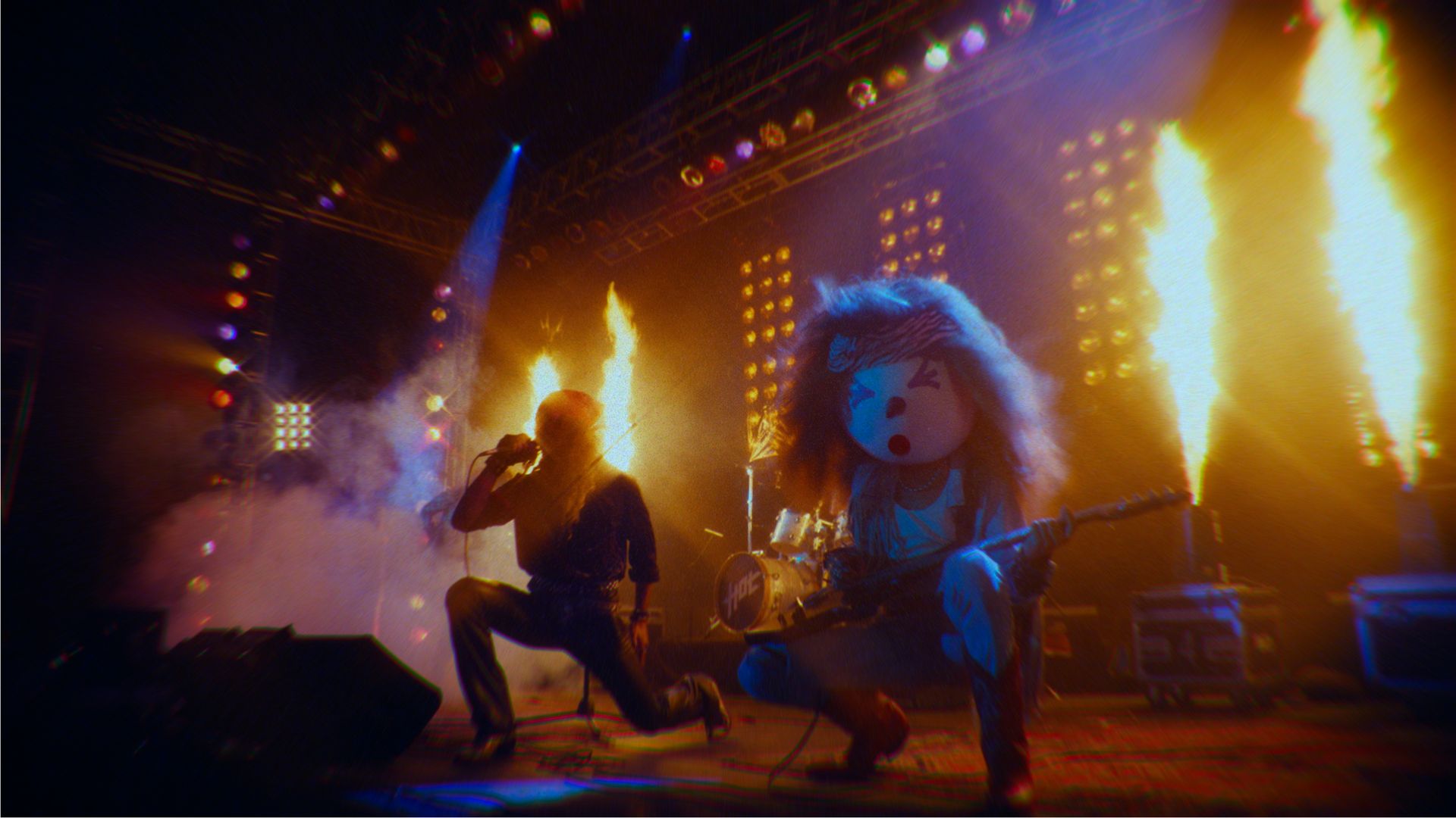 A live rock concert stage with fiery pyrotechnics; a singer kneels with a microphone and a guitarist wearing a large cartoonish head with wild hair and a headband performs amid bright yellow-orange flames.