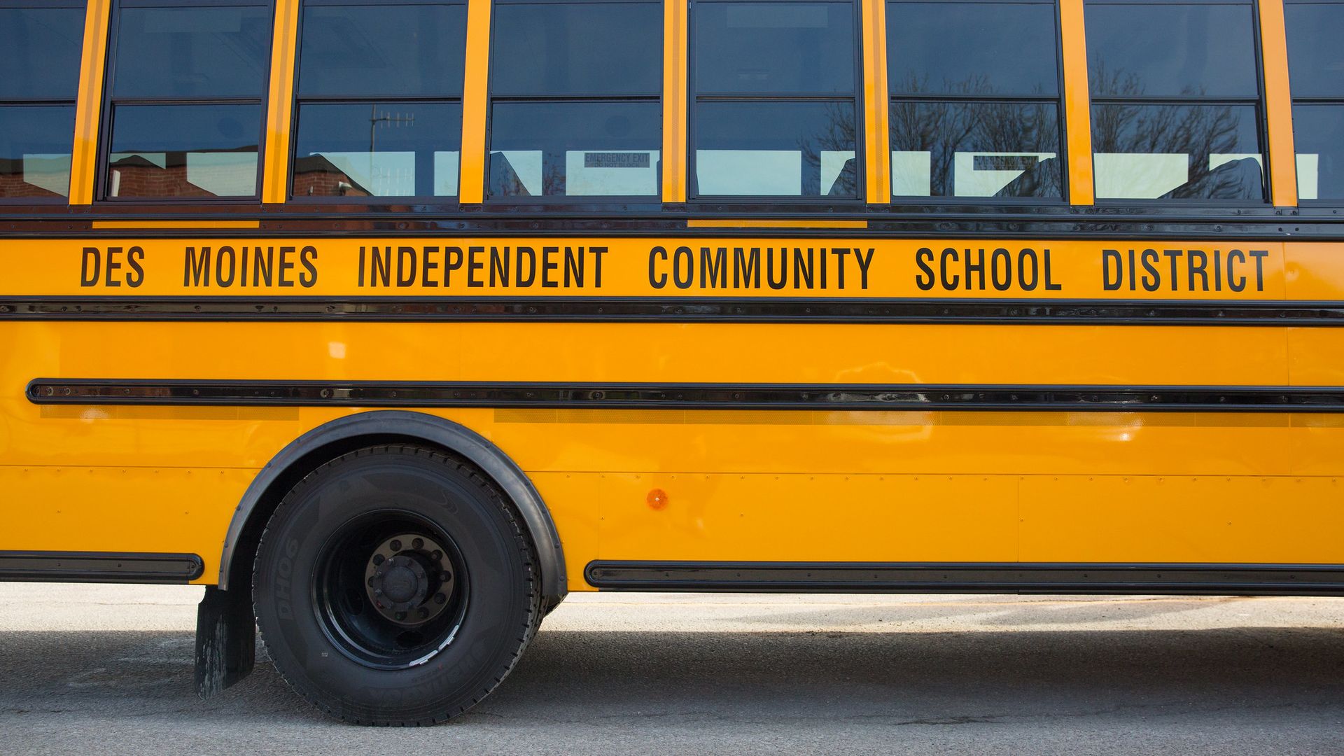 Side view of a yellow school bus with black lettering that reads "Des Moines Independent Community School District" and visible black tire and emergency exit signs.