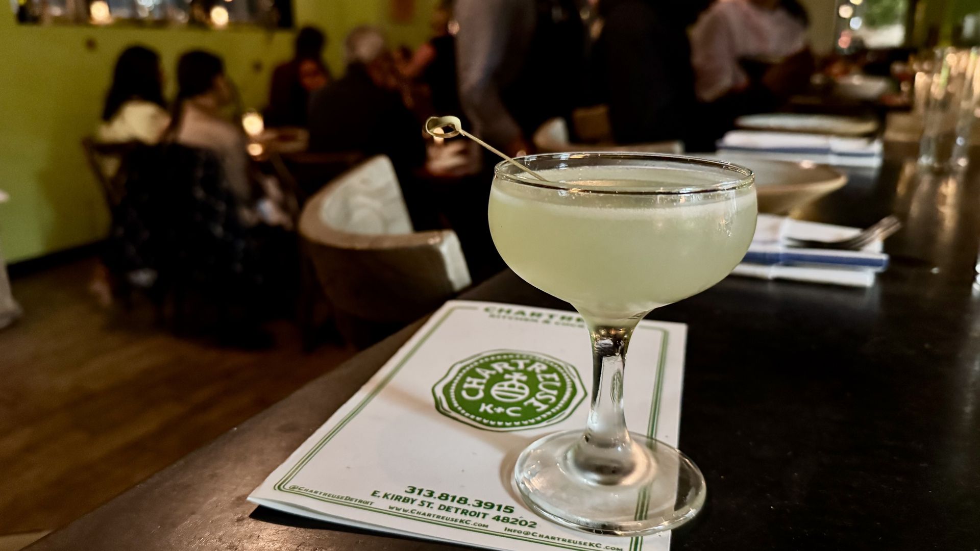 The pale green cocktail sits on a bar. In the background you see a green wall and patrons and a green menu