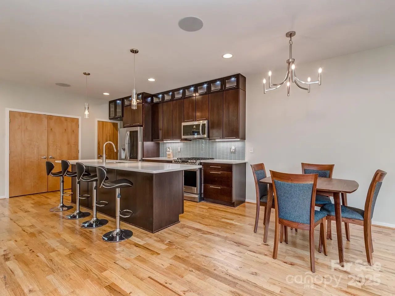 Modern kitchen with dark wood cabinets, stainless steel appliances, and a white island with four black bar stools on light wooden flooring, next to a dining table with four blue-upholstered chairs.