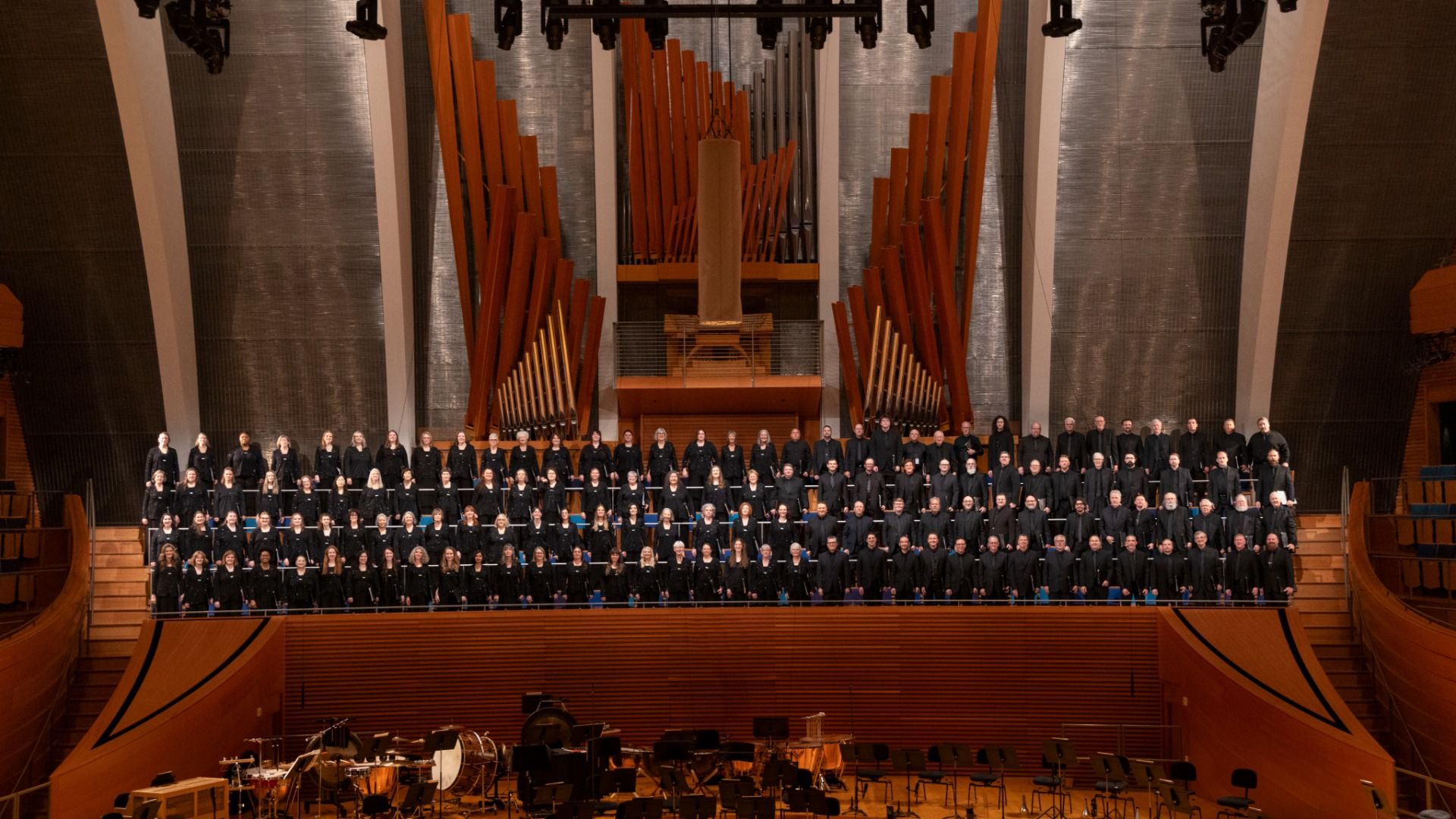 A large choir in black robes stands on a balcony in a concert hall, with tall orange organ pipes rising behind them and a stage filled with instruments in front.
