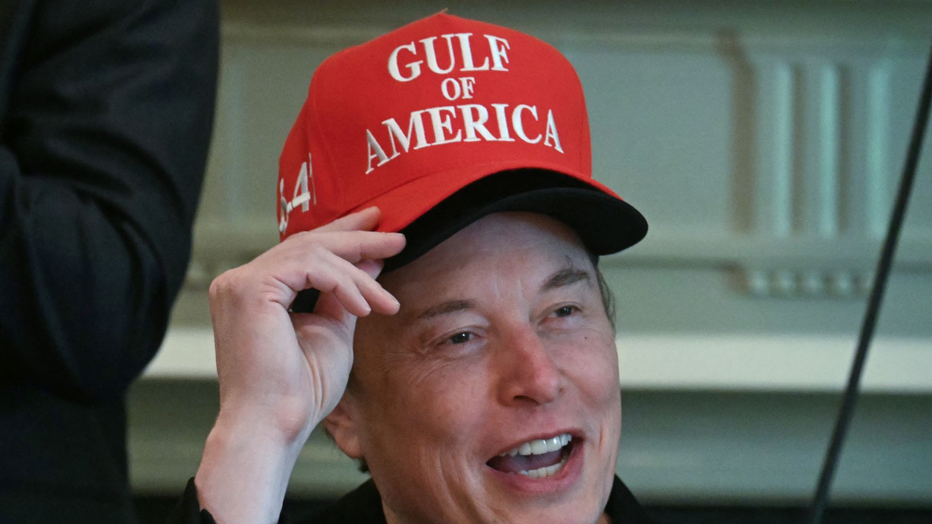 Elon Musk wearing two hats, the top, red one saying "Gulf of America" 