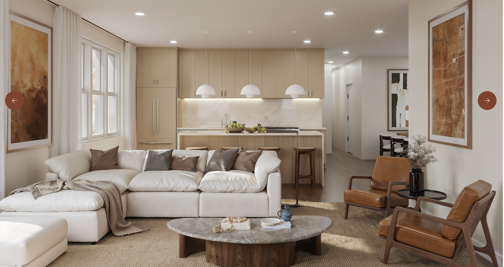Open-plan living area with a white sectional sofa, round marble coffee table, and tan leather chairs. In the back, a light-wood kitchen with pendant lights and marble backsplash, plus framed art.