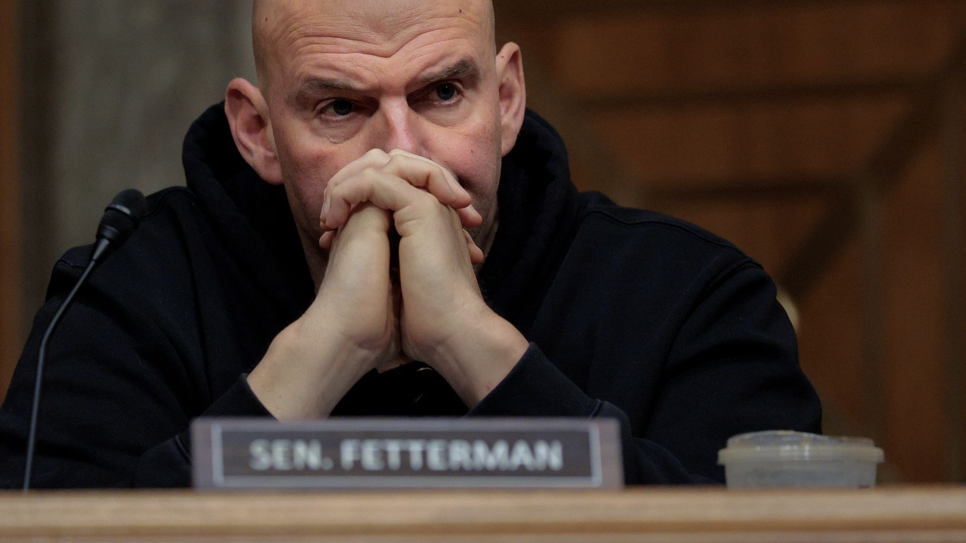 Close-up of Sen. Fetterman in a dark hoodie, hands folded under his chin at a desk. A microphone, a "SEN. FETTERMAN" nameplate, and a small cup are visible.