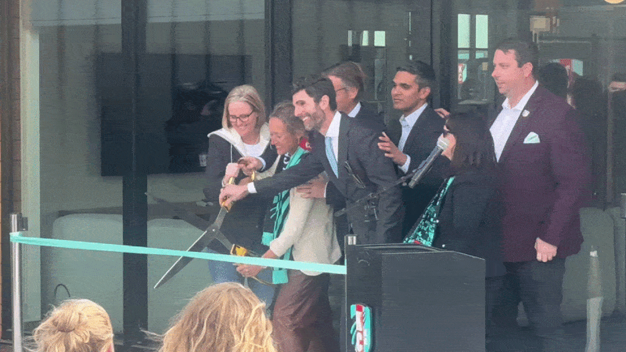 Group of people in business attire cutting a teal ribbon at a ceremony with a microphone and audience visible, indicating an official opening event.