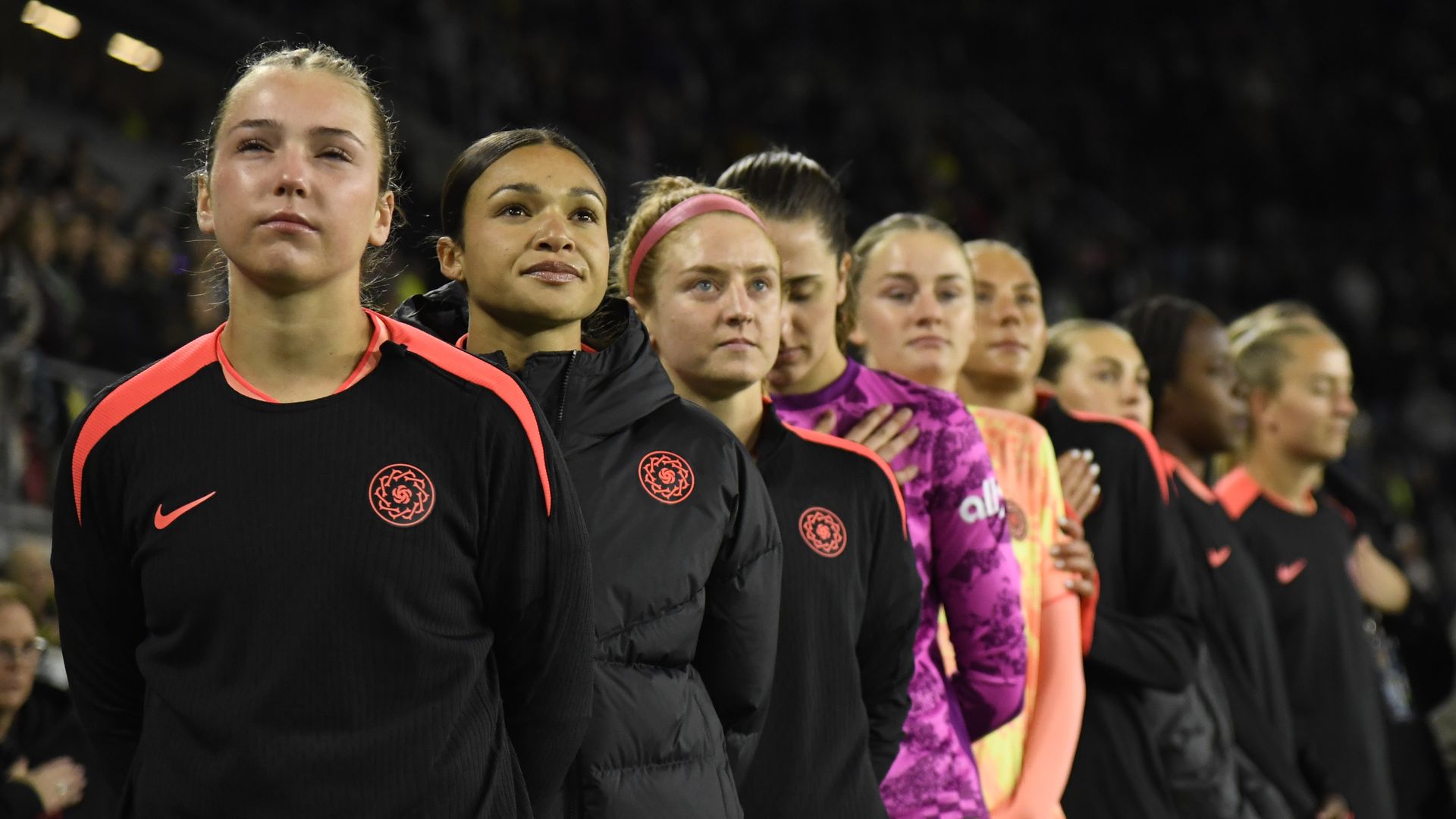 Line of female soccer players standing in a stadium lineup; foreground player wears black with red trim and crest, others in jackets or neon pink/purple jerseys behind.
