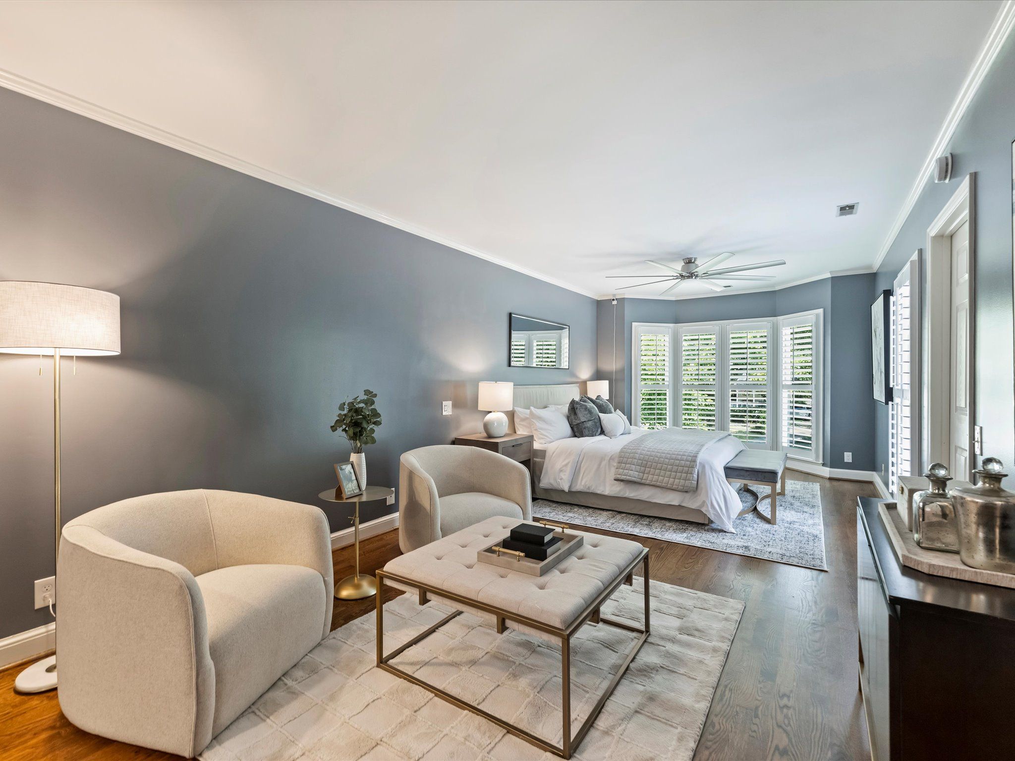 Modern bedroom with gray walls, wood floor, white bed, beige armchairs, tufted coffee table, standing lamp, bay window with white shutters, and ceiling fan.